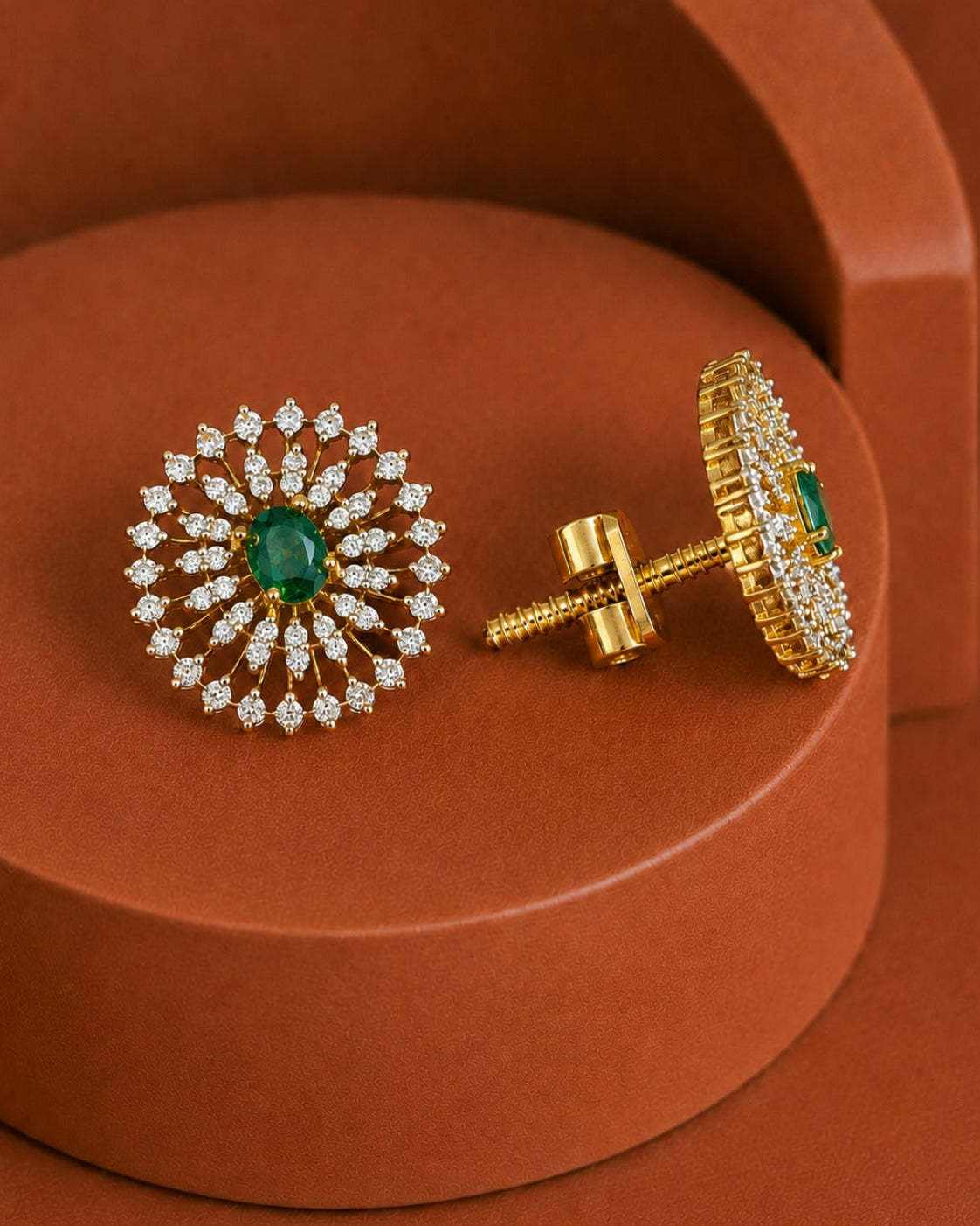 A pair of gold post earrings with a circular sunburst design, featuring a central green emerald stone and surrounding rows of small clear stones, displayed on a textured terracotta background.