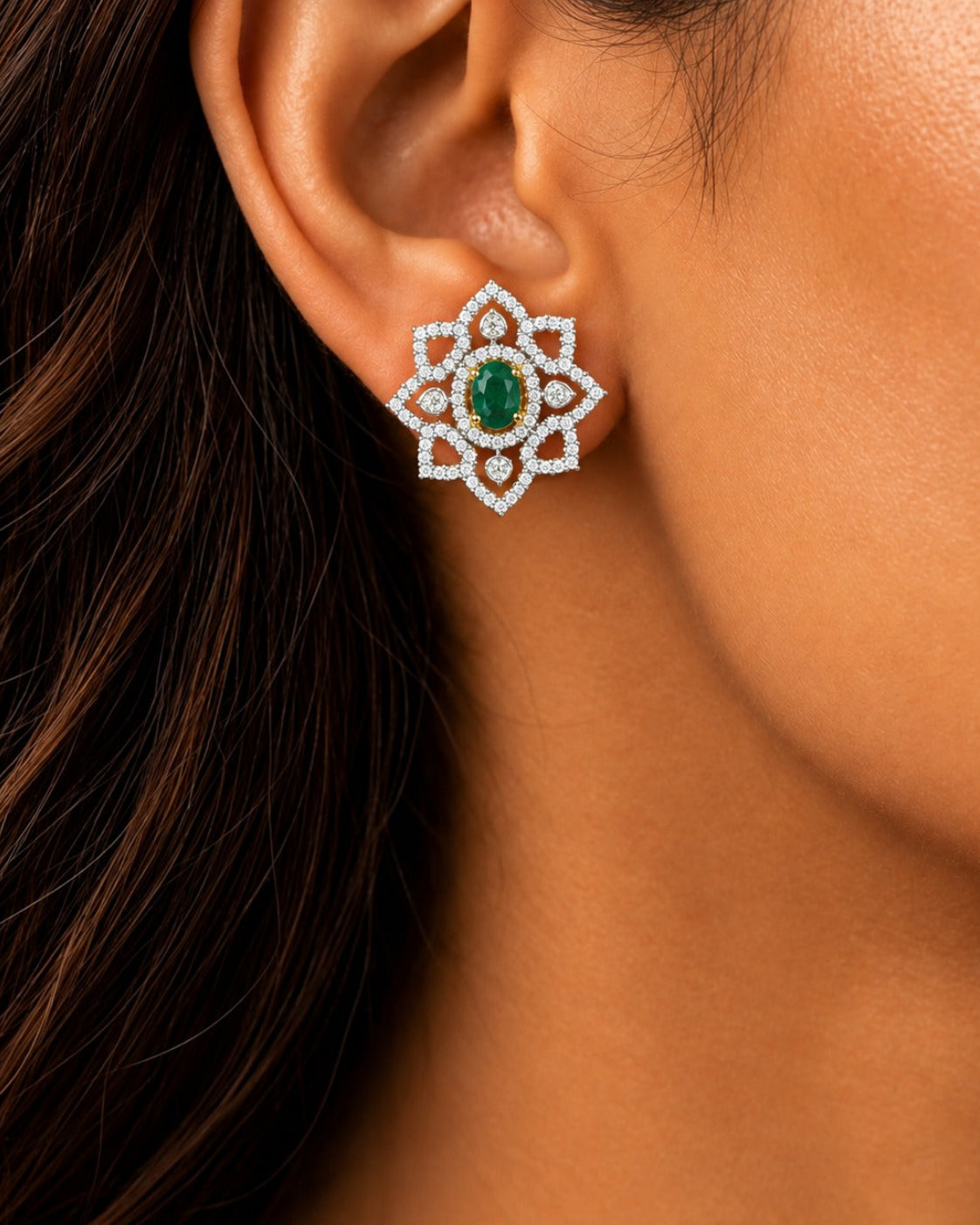 A close-up of a woman's ear wearing a floral-shaped stud earring with a green oval center stone and a surrounding border of small clear stones.