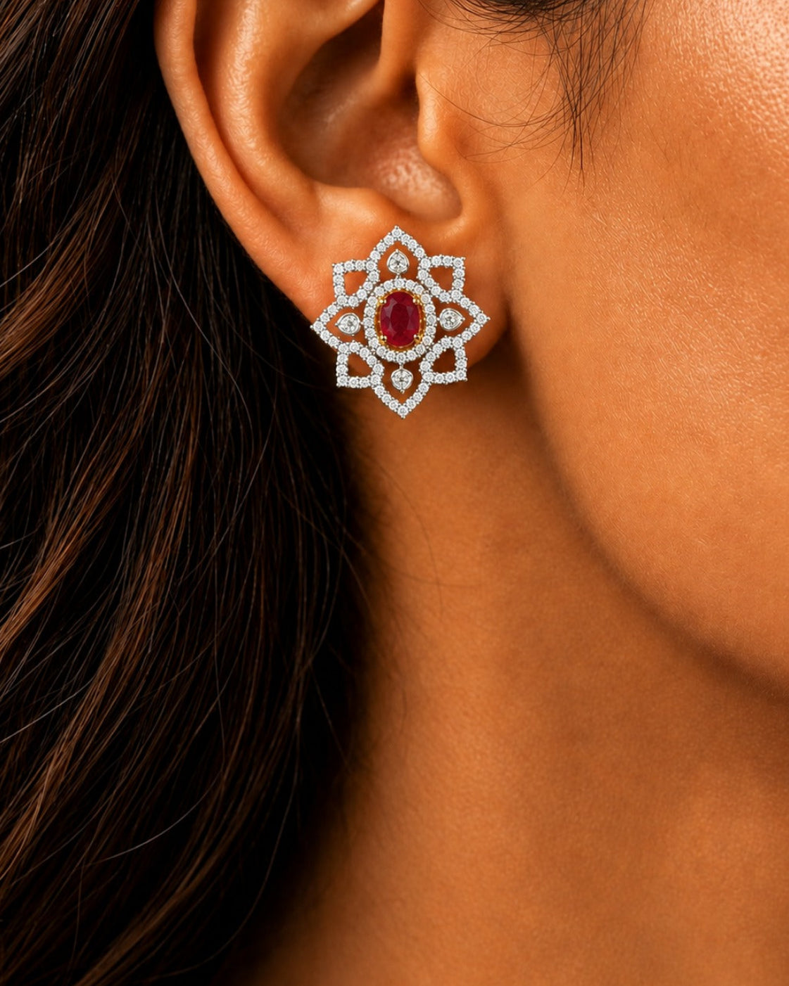 A close-up of a woman's ear wearing a floral stud earring with a central oval red gemstone surrounded by a halo of small clear stones in a silver-toned setting.