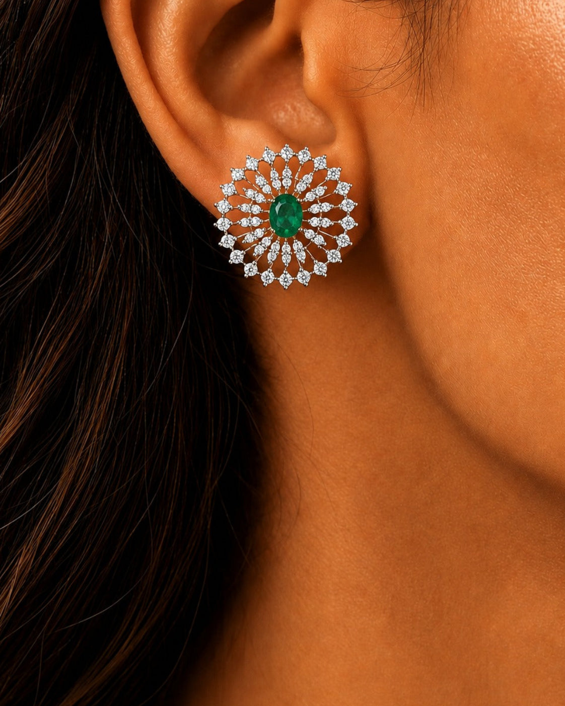 A close-up of a woman's ear wearing a circular stud earring with a green emerald center and surrounding silver-toned and clear stones.