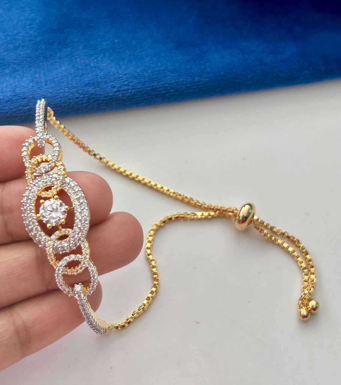 A close-up of a gold and silver-toned bracelet with a central clear gemstone, held by a hand against a white surface with a blue fabric background.
