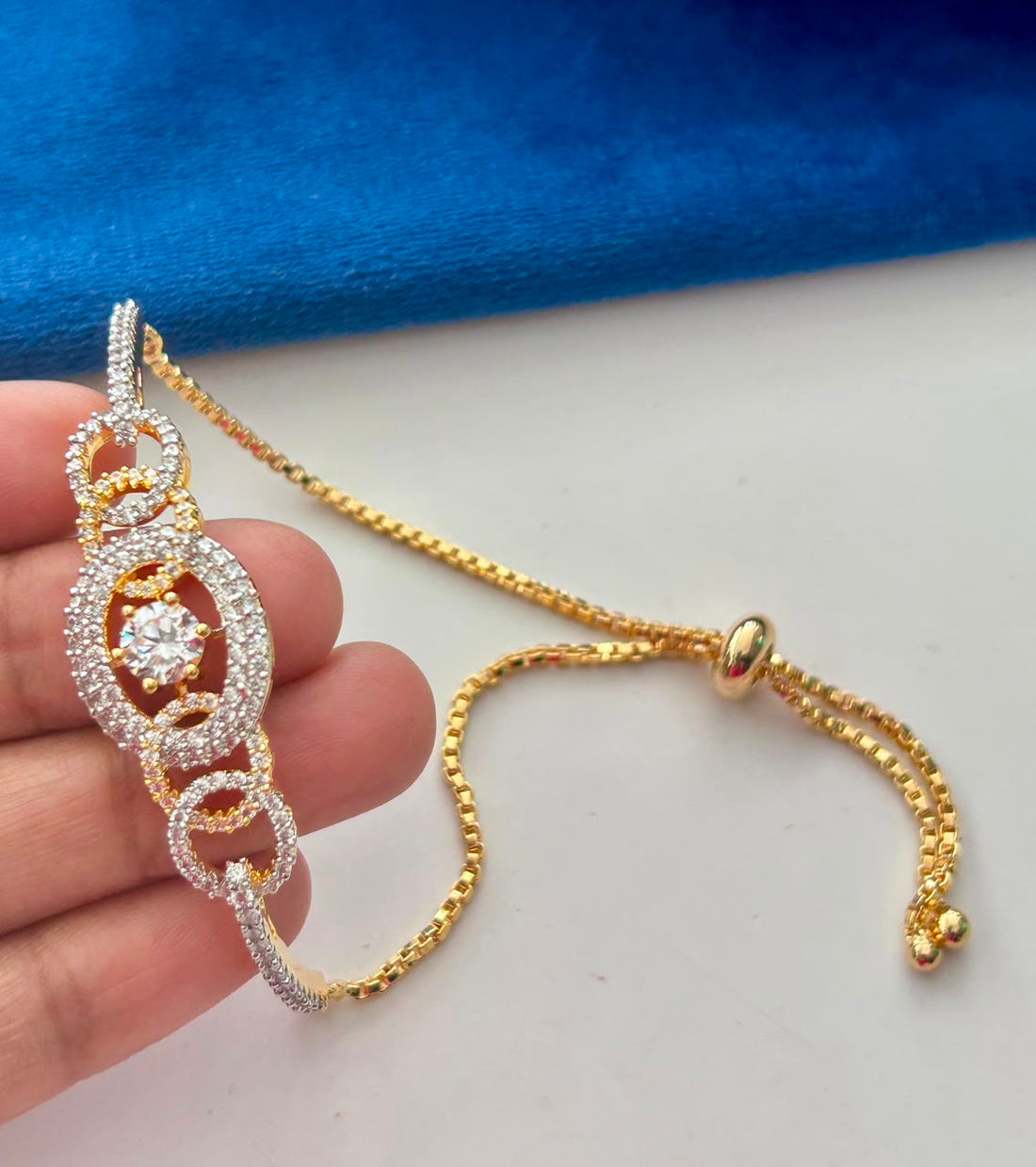 A close-up of a gold and silver-toned bracelet with a central clear gemstone, held by a hand against a white surface with a blue fabric background.