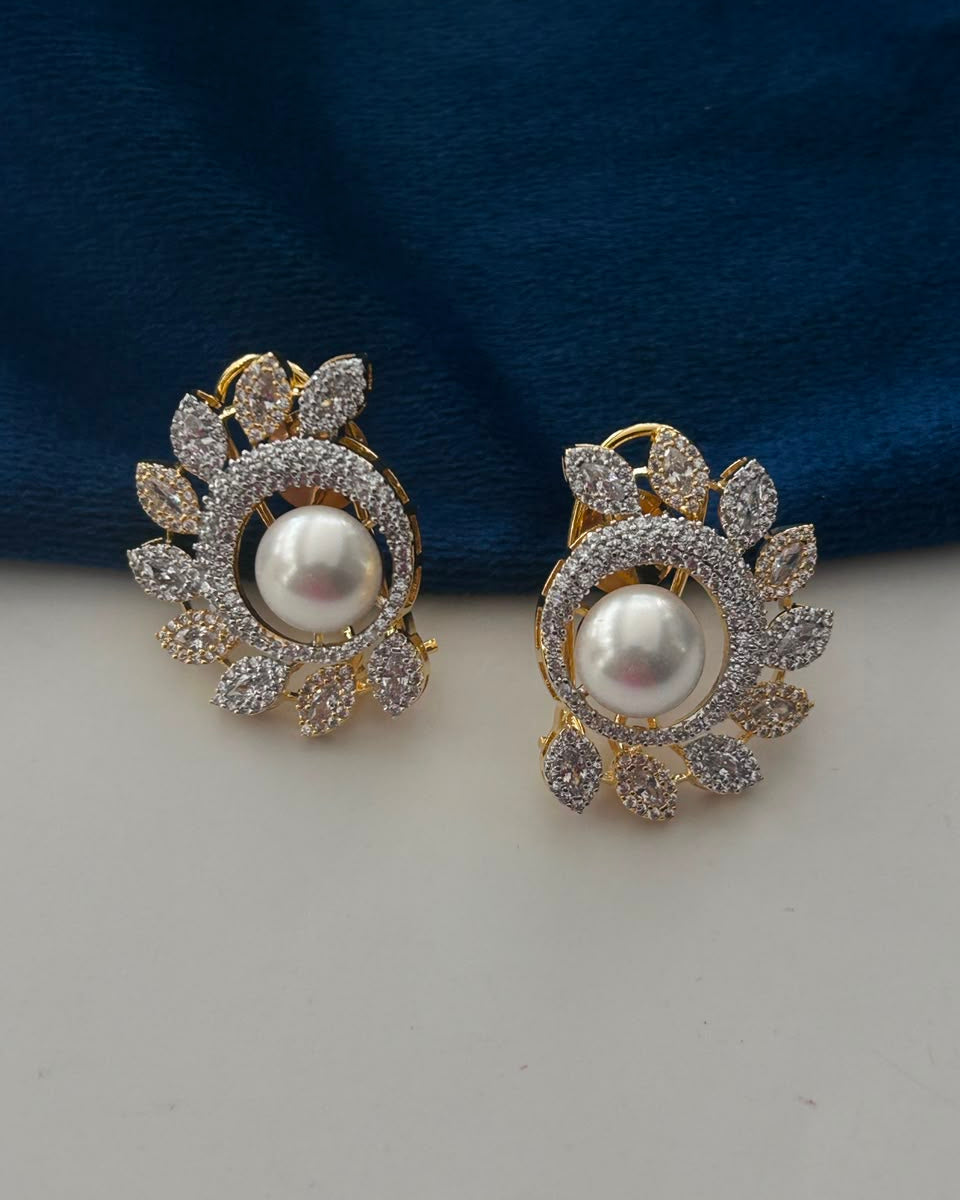 A pair of gold-toned clip-on earrings featuring a central white pearl surrounded by pavé-set clear stones in a leafy design, displayed on a white surface with a dark blue fabric background.
