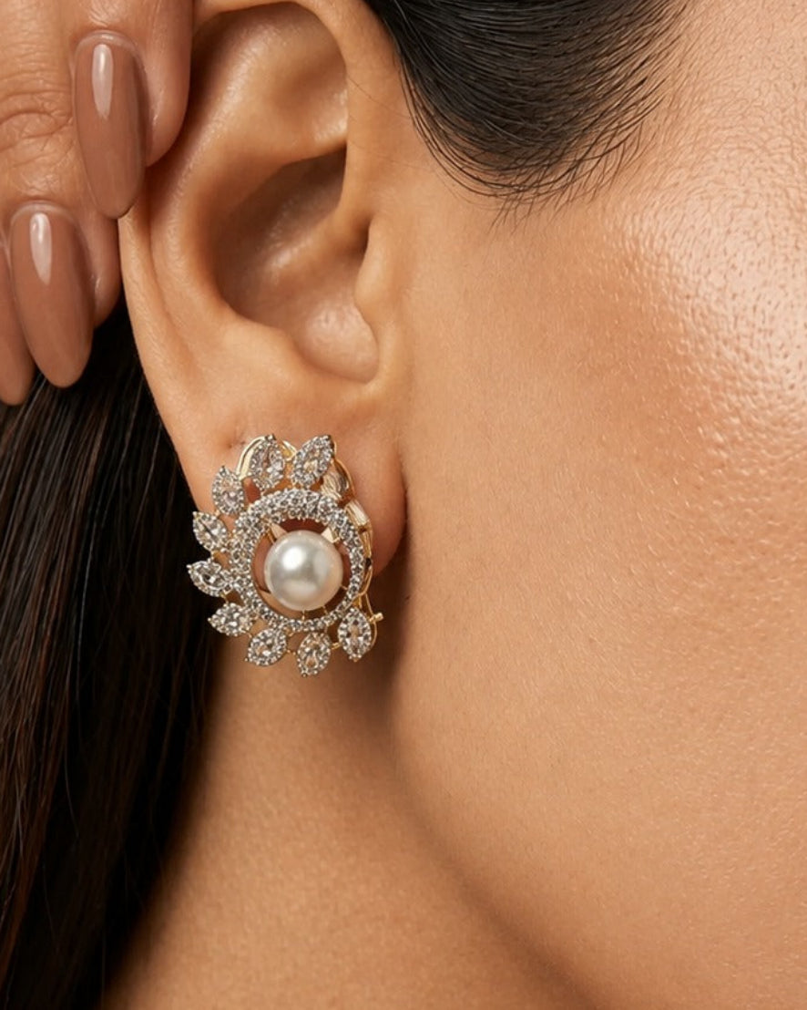 A close-up of a woman's ear wearing a floral-shaped stud earring with a central white pearl and surrounding clear stones in a gold-tone setting.
