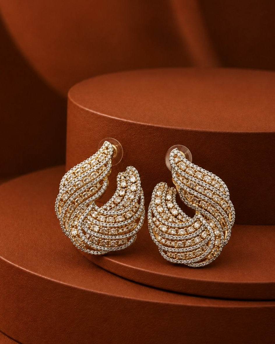 A pair of gold-toned hoop earrings covered in rows of small clear stones, displayed on a round brown pedestal against a matching brown background.
