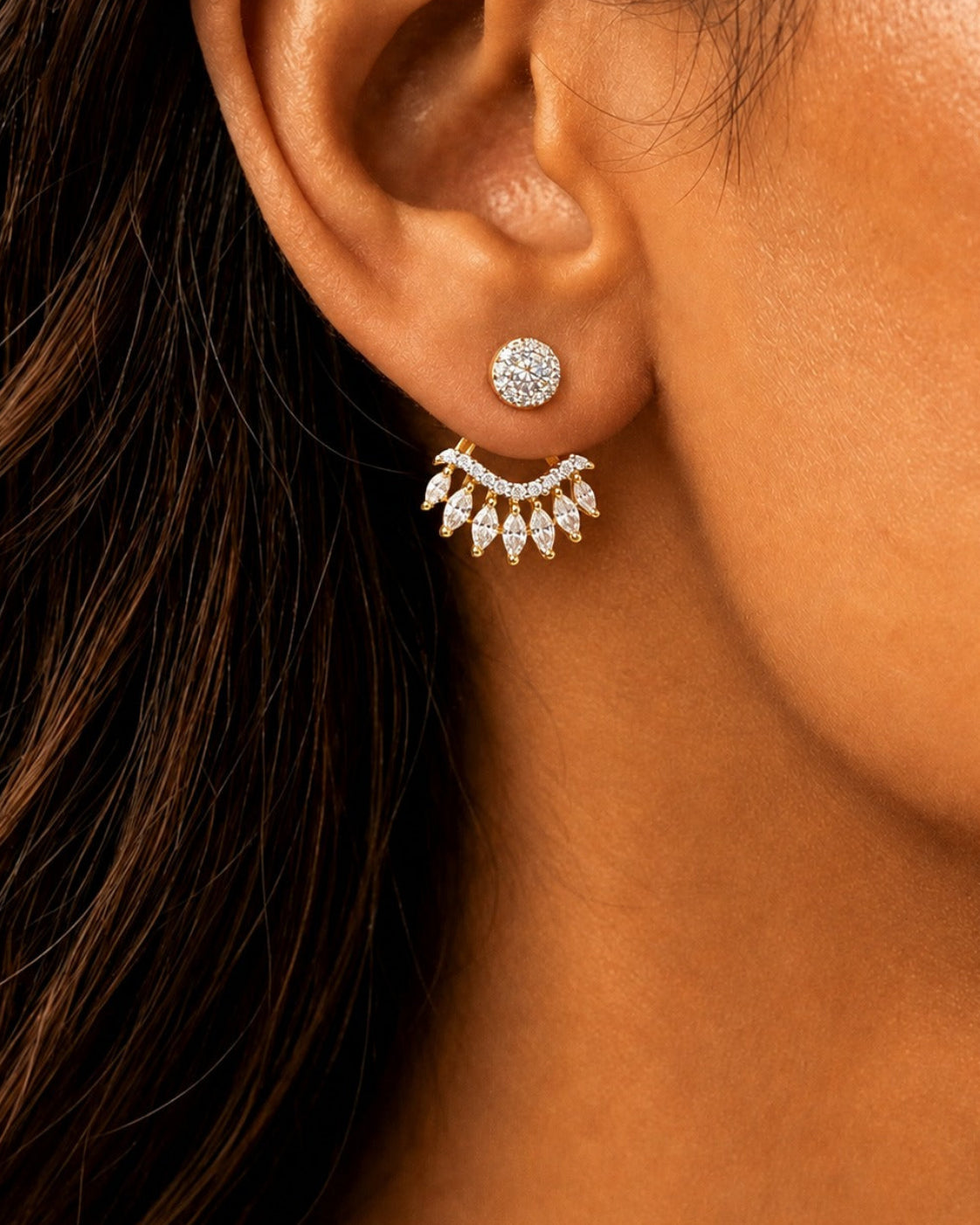 A close-up of a model's ear wearing a gold stud earring with a round pavé top and a dangling leaf-shaped pavé chandelier drop.
