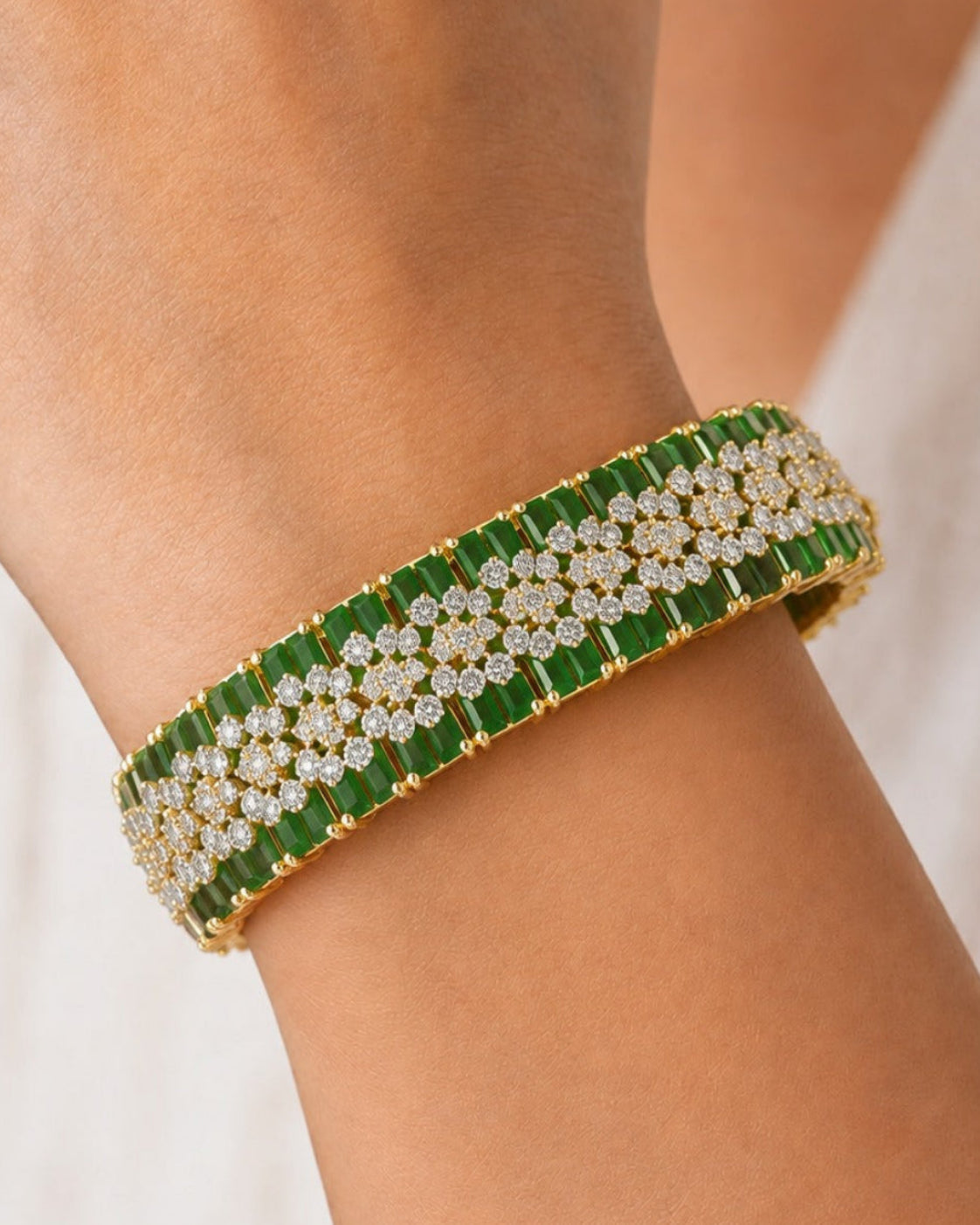 A close-up of a gold-plated cuff bracelet with green rectangular stones and rows of clear pavé diamonds, worn on a person's wrist.