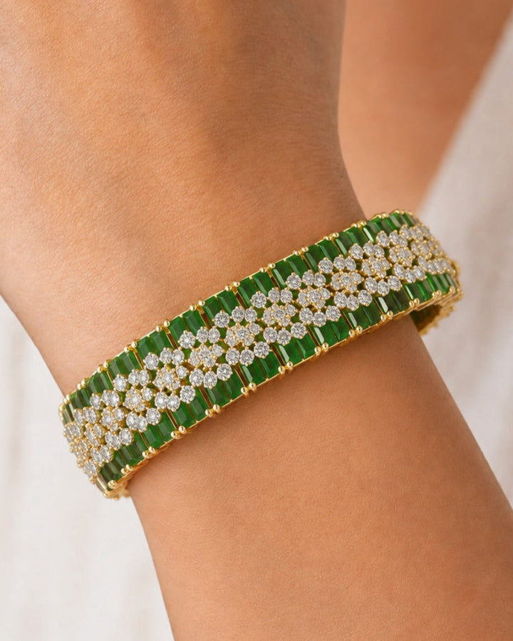 A close-up of a gold-plated cuff bracelet with green rectangular stones and rows of clear pavé diamonds, worn on a person's wrist.