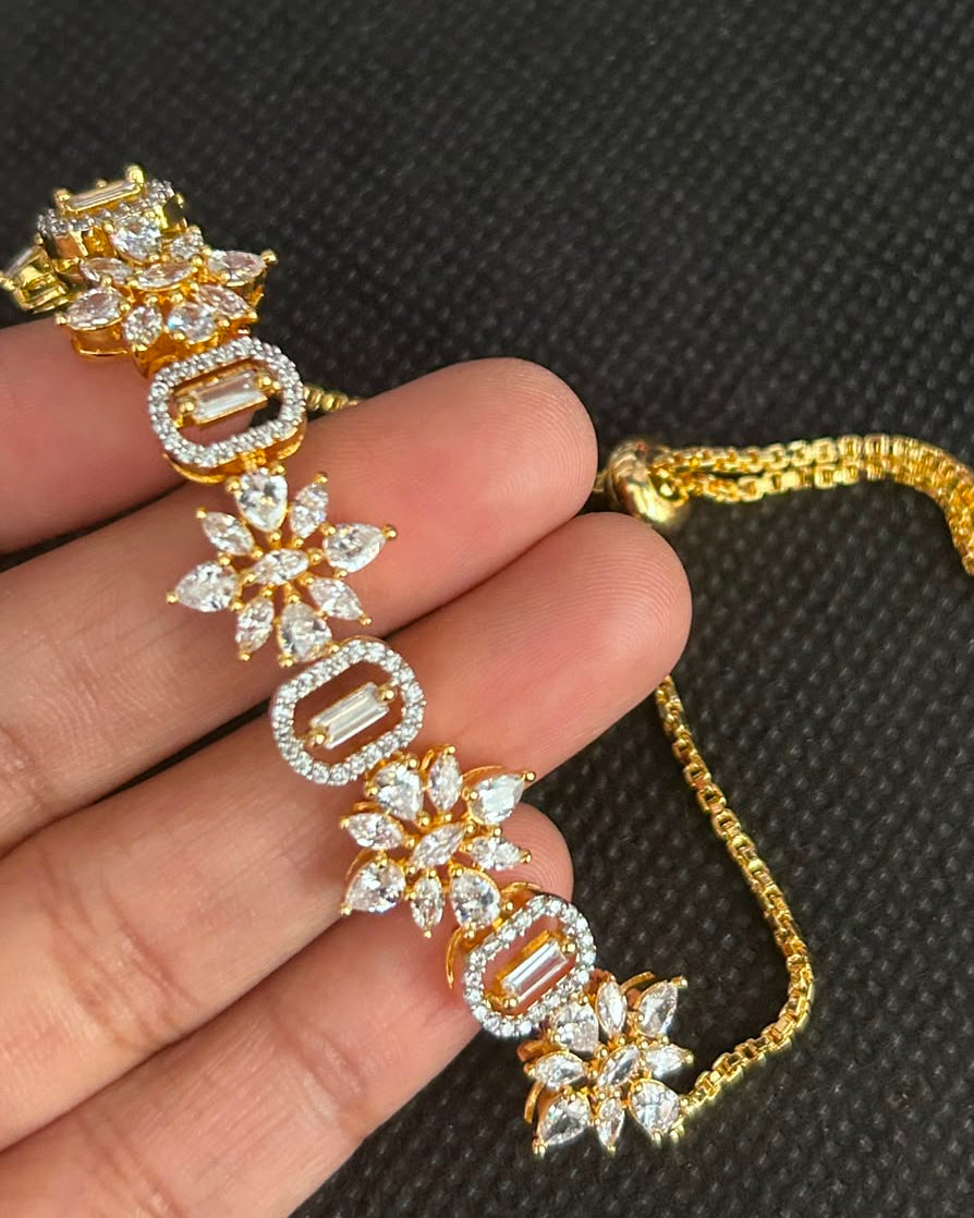 A gold bracelet with clear pavé-set stones in floral and rectangular motifs, held on a person's fingers against a dark textured background.