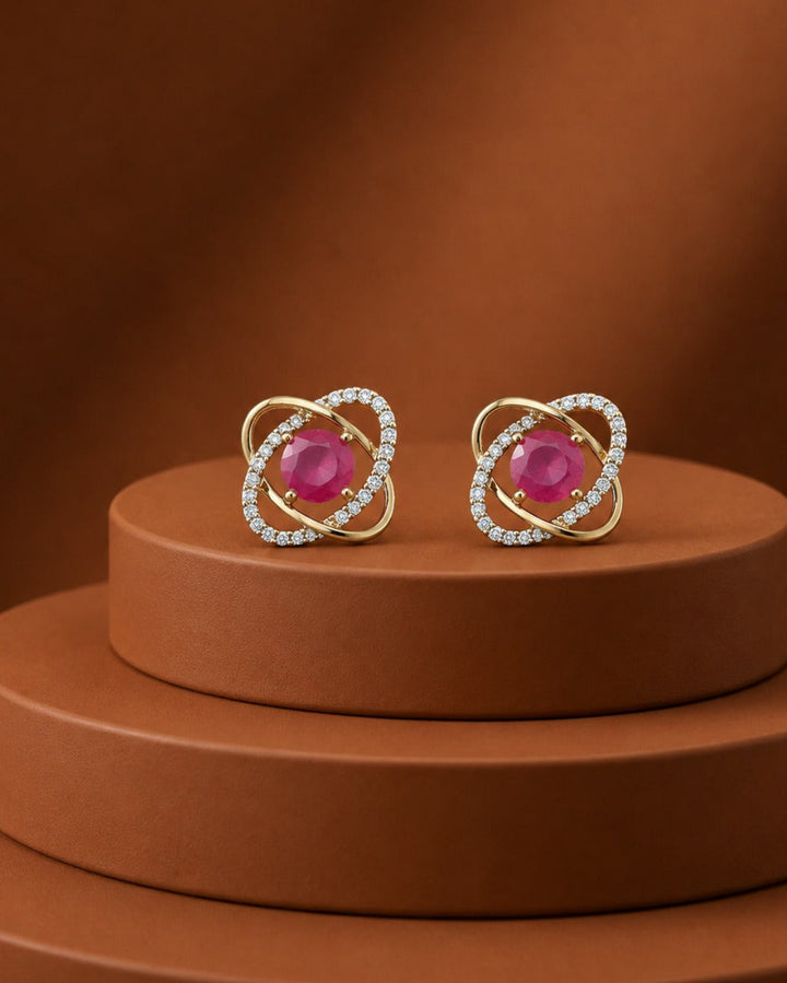 A pair of gold-toned stud earrings with oval pink gemstones and surrounding rows of small clear stones, displayed on a brown tiered pedestal against a matching brown background.