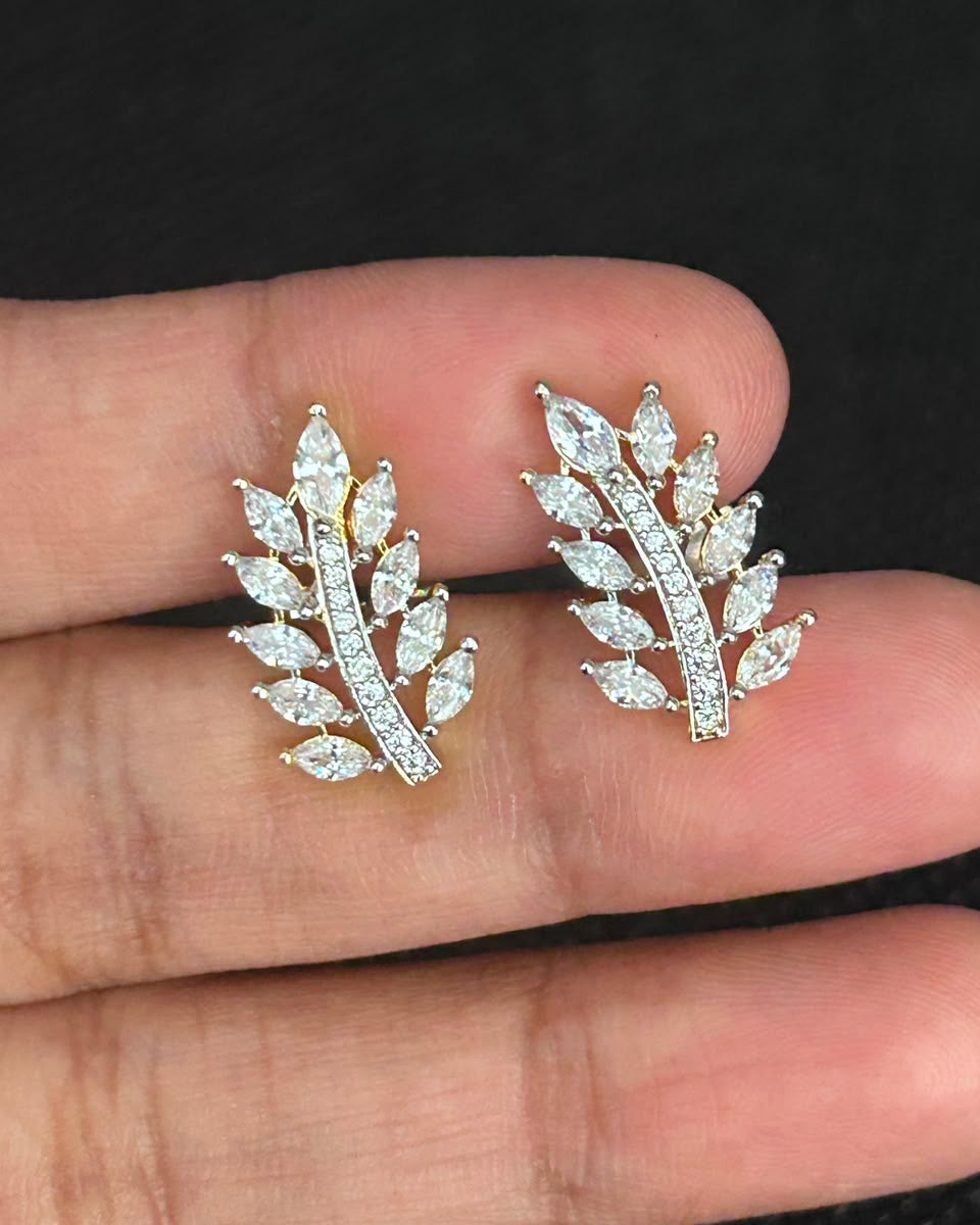 A pair of gold leaf-shaped stud earrings set with clear marquise and round stones, shown resting on a person's fingers against a dark background.