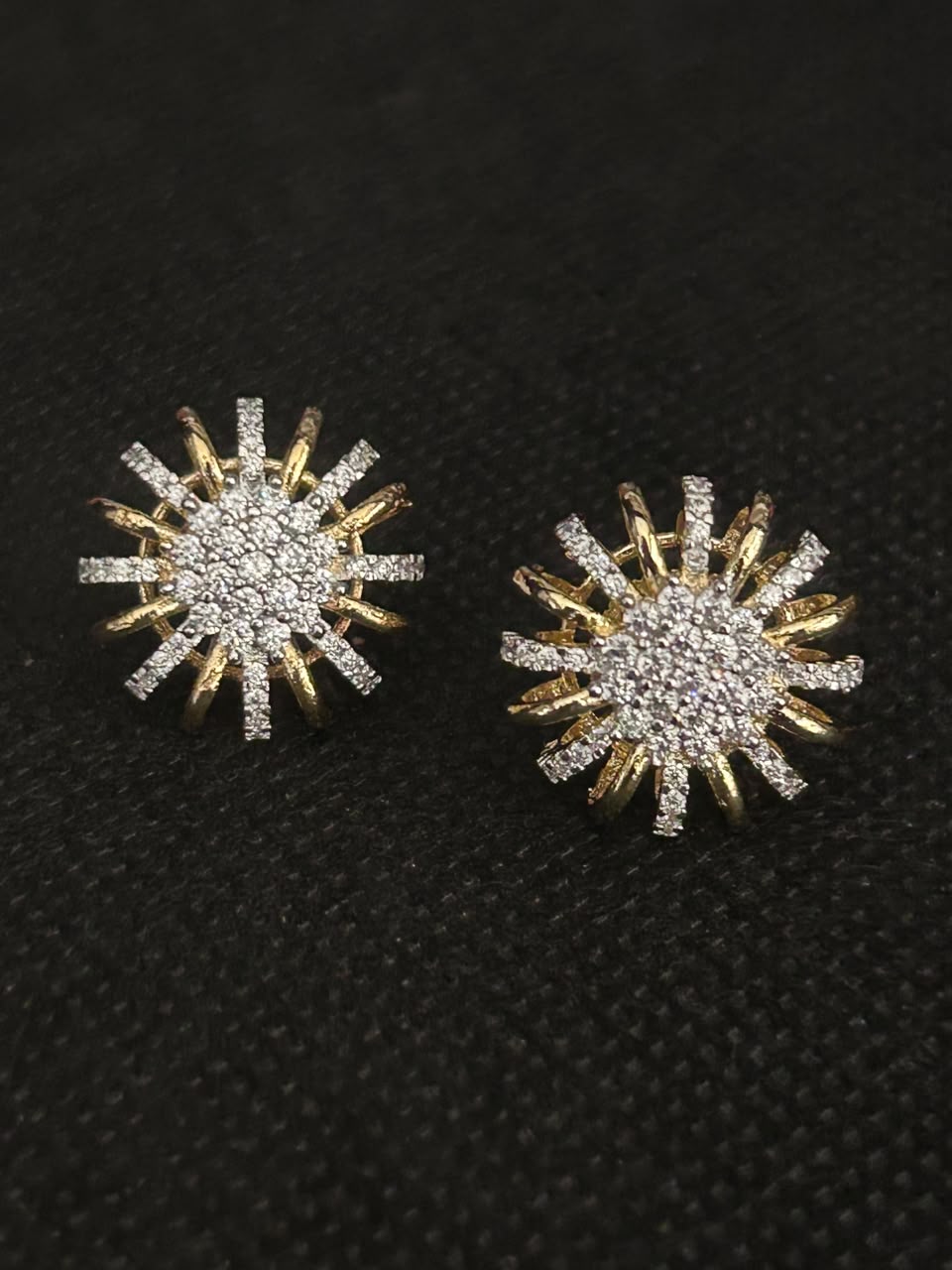 A pair of gold and silver starburst stud earrings paved with clear stones, displayed on a textured black background.