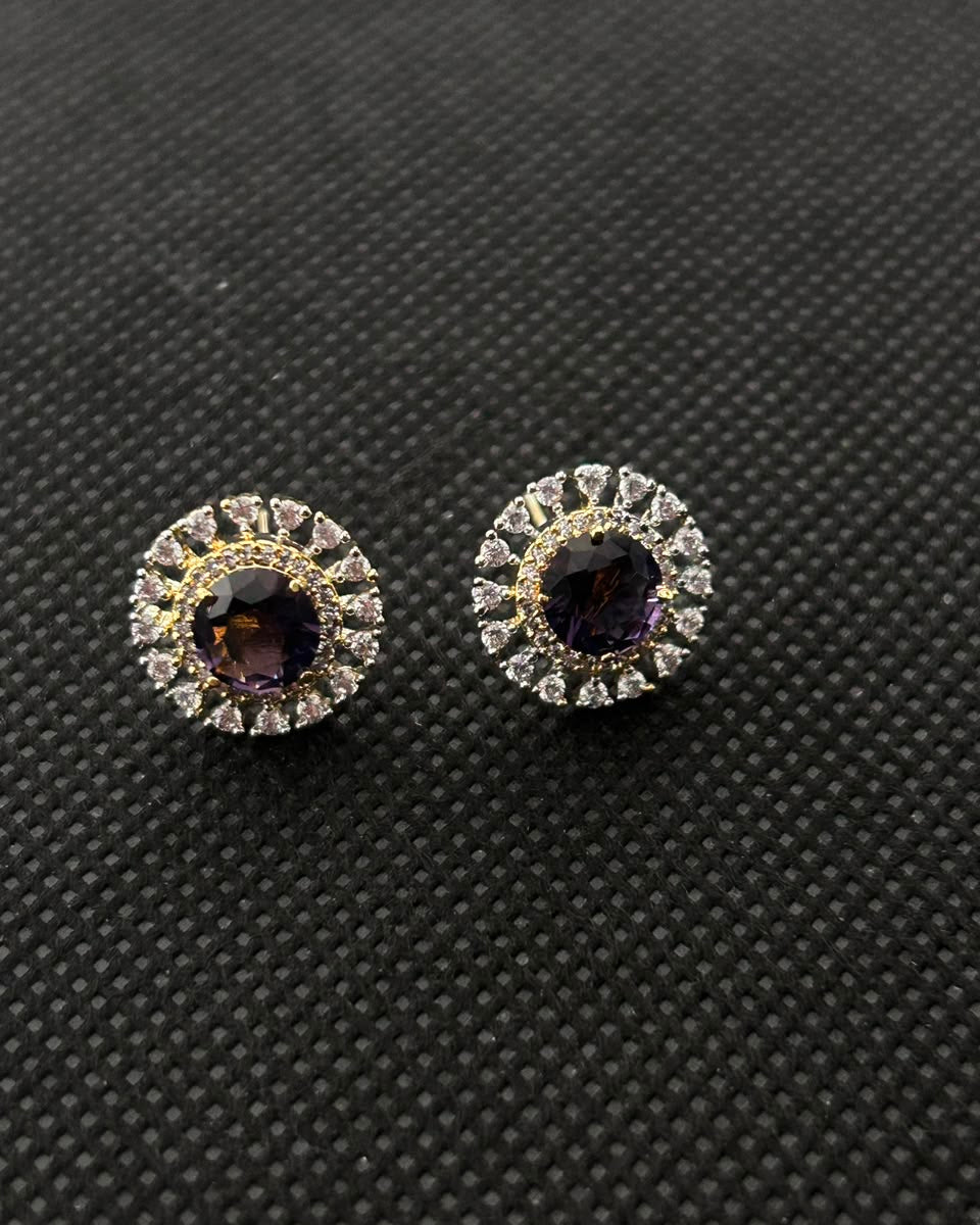 A pair of round stud earrings with dark purple center stones surrounded by clear halo crystals and gold accents, displayed on a textured black background.