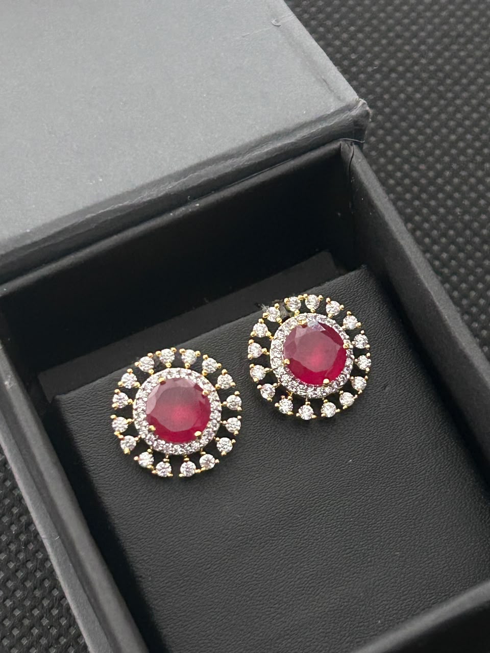 A pair of round stud earrings with vibrant red center stones surrounded by small clear stones in a gold-tone halo setting, displayed inside an open black jewelry box.