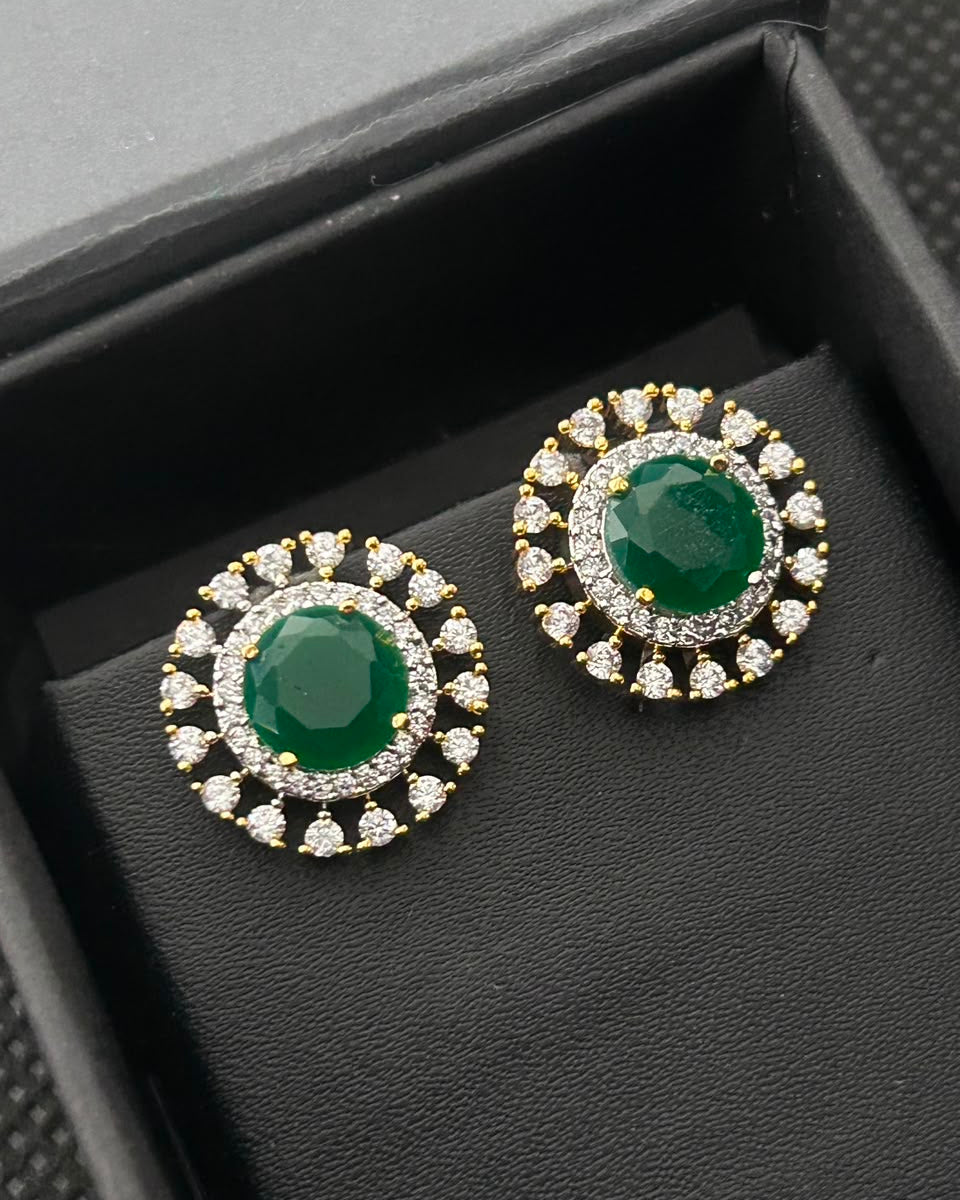 A pair of gold-plated stud earrings with oval green gemstones surrounded by clear cubic zirconia, displayed in a black jewelry box.