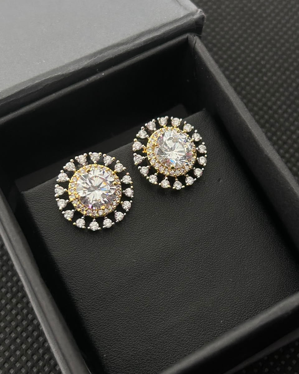 A pair of round stud earrings with clear gemstones in a gold and silver-tone halo setting, displayed inside a black jewelry box.