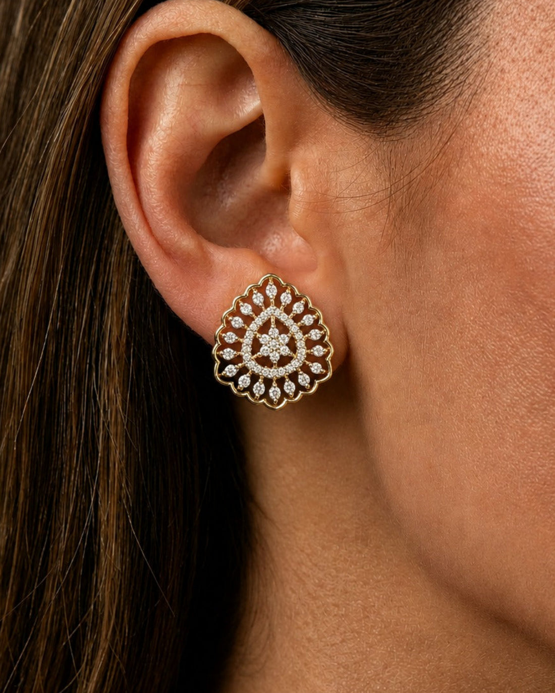 A close-up of a woman's ear wearing a detailed gold-toned stud earring with small clear stones arranged in a circular pattern.
