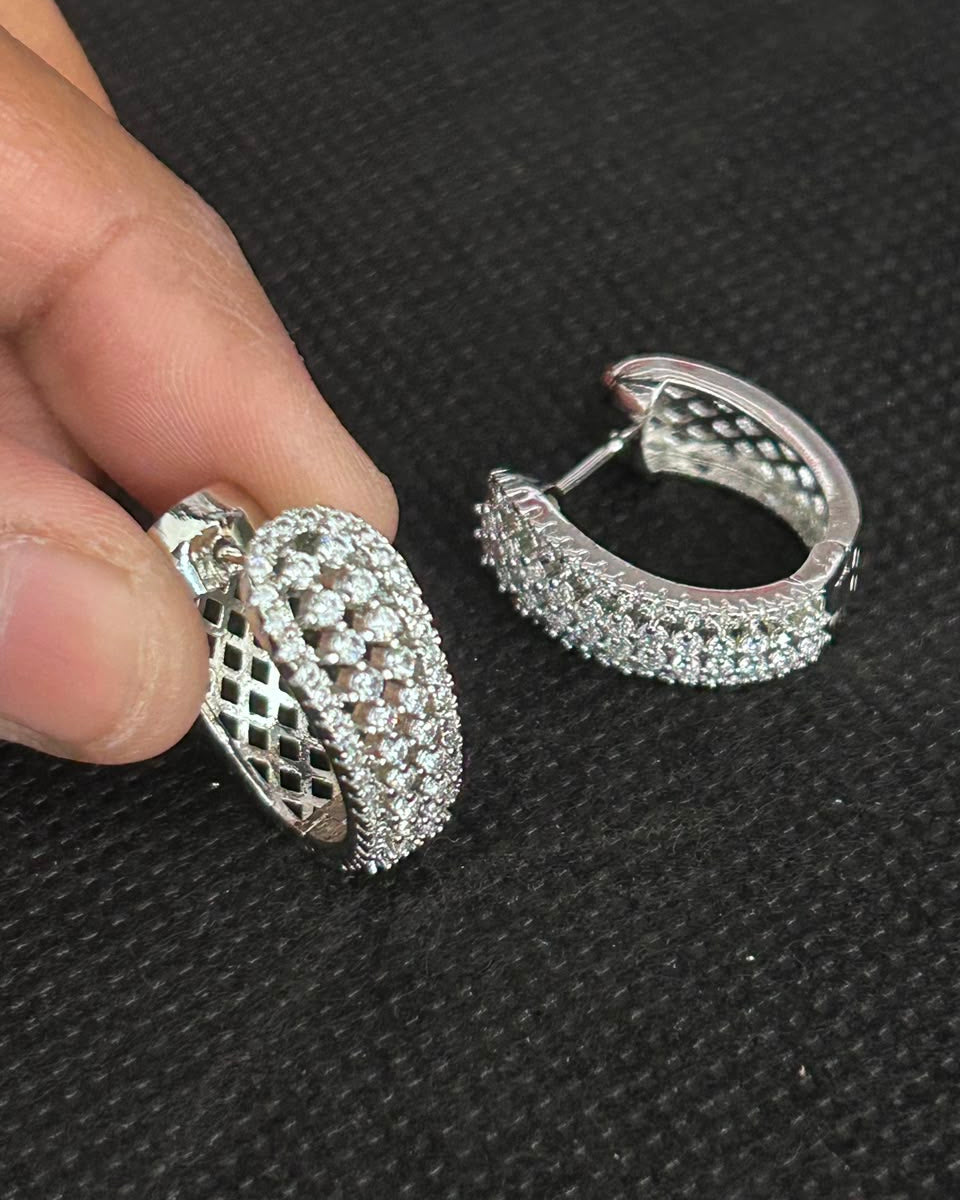 A pair of silver hoop earrings paved with clear stones, with one earring held by a hand and the other resting on a textured black background.