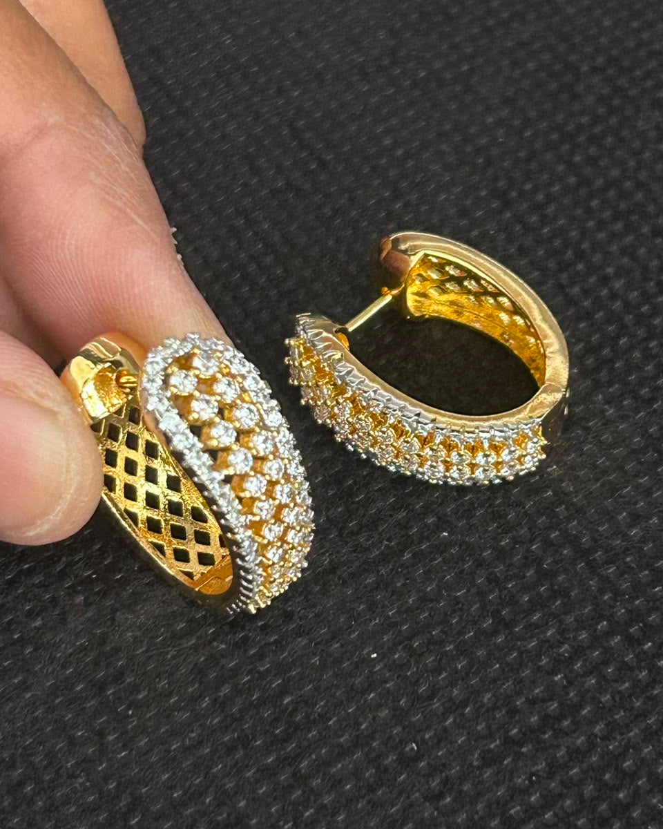 A pair of gold huggie hoop earrings with pave-set clear stones and a textured gold lattice band, shown with a person's fingers holding one earring against a black textured background.