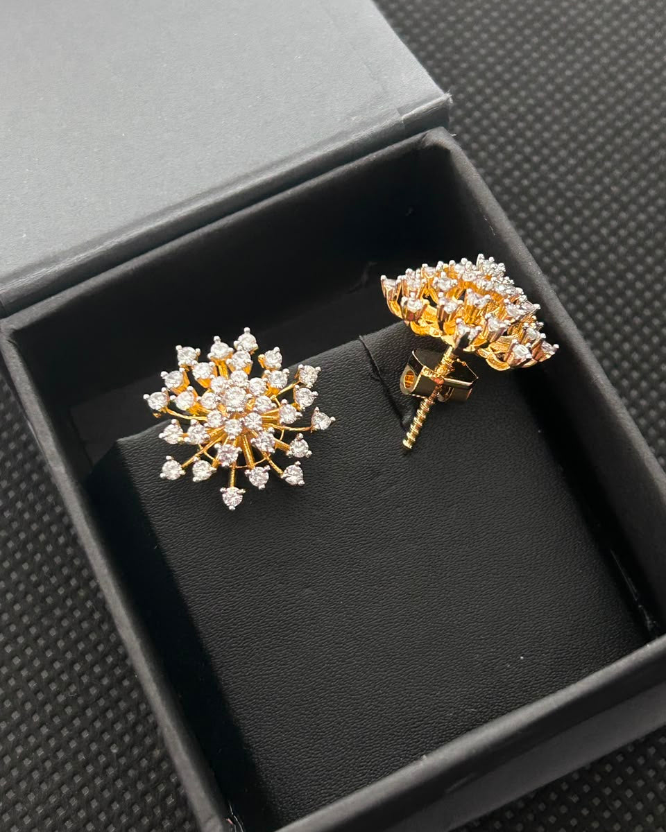 A pair of gold flower-shaped stud earrings set with clear gemstones, displayed inside an open black jewelry box on a textured dark background.