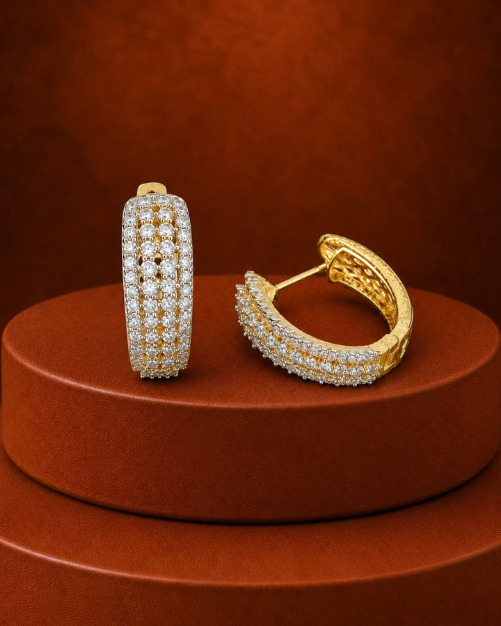 A pair of gold huggie hoop earrings paved with small clear stones, displayed on a round brown pedestal against a matching brown background.