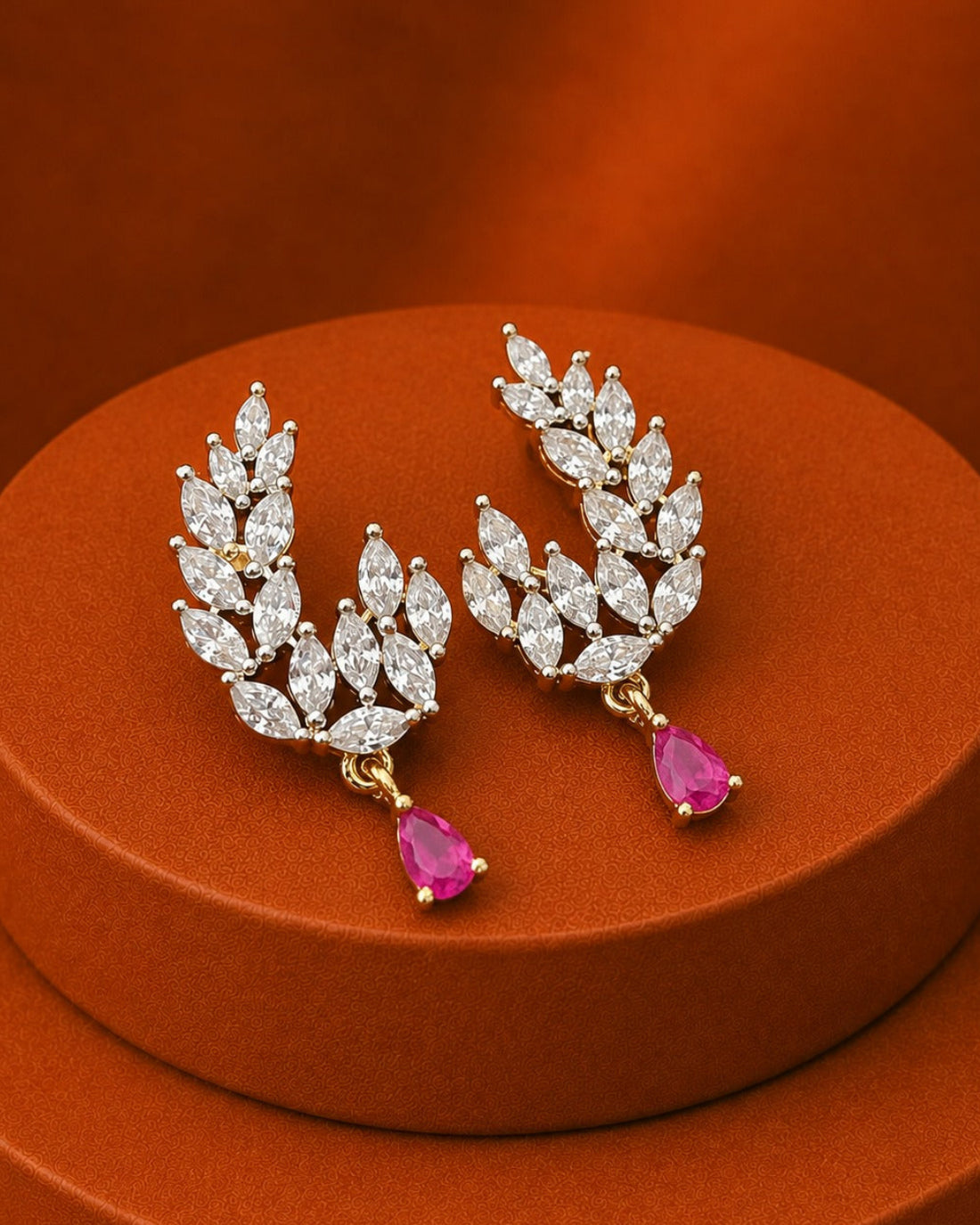 A pair of gold leaf-shaped drop earrings set with clear marquise and oval stones, each ending in a pink teardrop gemstone, displayed on a round orange pedestal against a matching orange background.