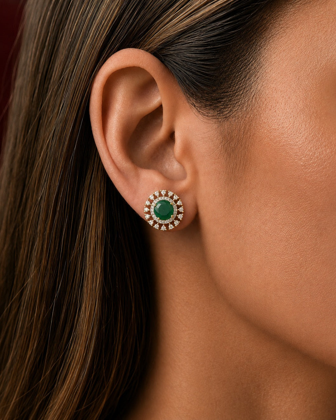 A close-up of a woman's ear wearing a round stud earring with a green center stone and a halo of small clear stones in a gold-toned setting.