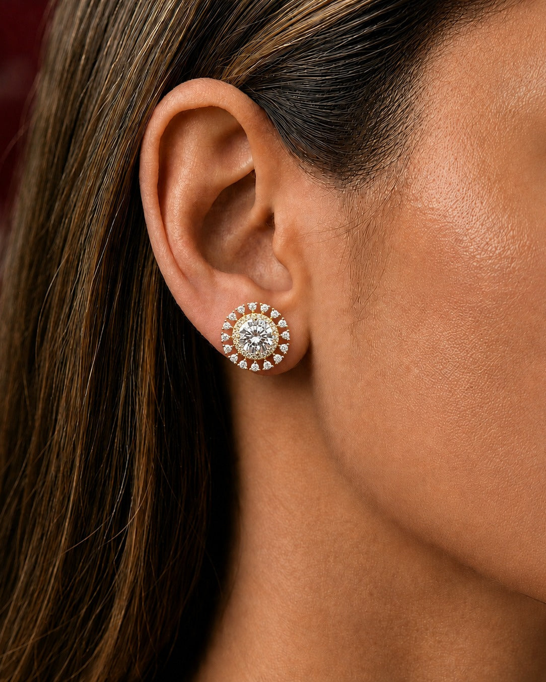 A close-up of a woman's ear wearing a round stud earring with a halo of small clear stones and a central clear gemstone in a gold-toned setting.