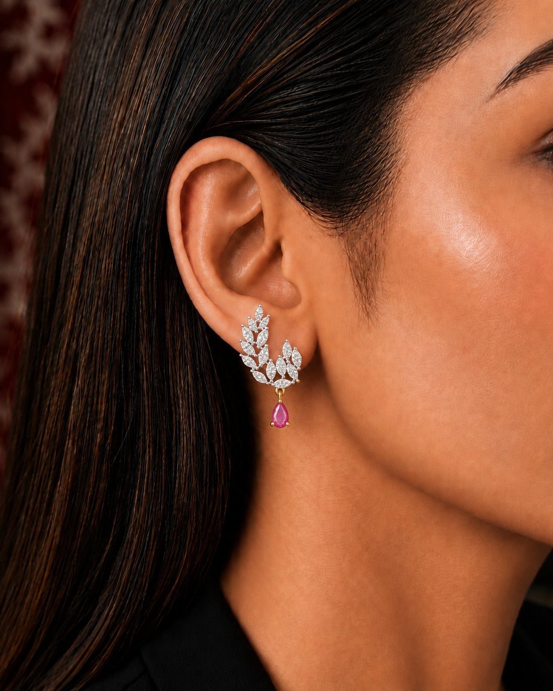 A close-up side profile of a woman wearing a silver-toned leaf-shaped drop earring with clear stones and a dangling pink teardrop gemstone.