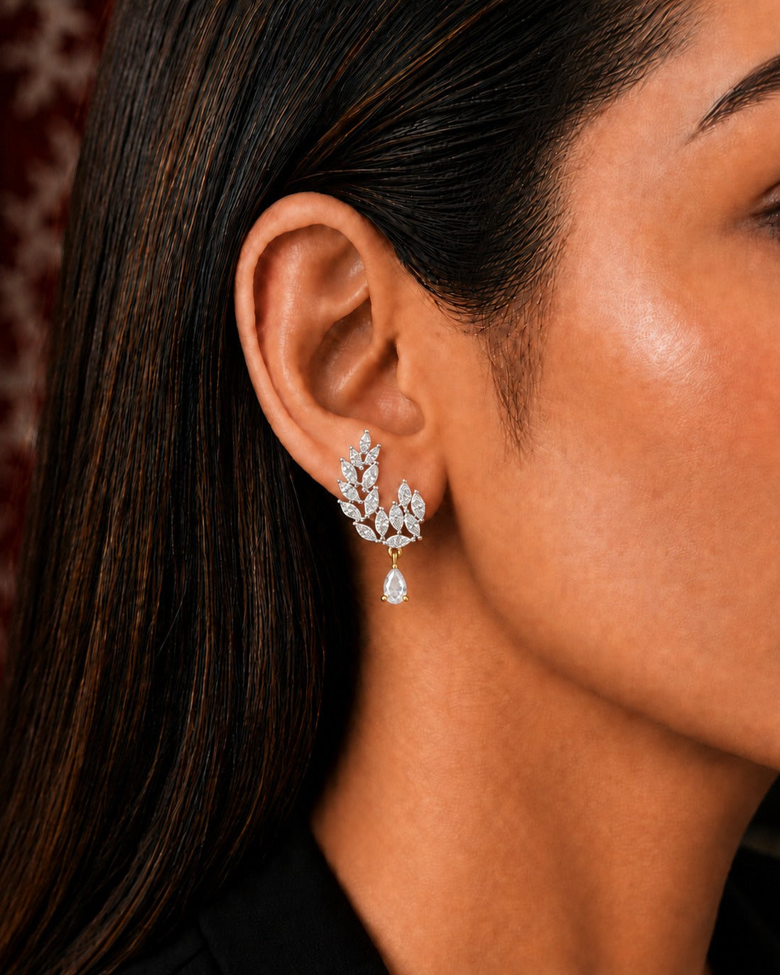A close-up of a model's ear wearing a delicate silver-toned leaf-shaped drop earring with a teardrop crystal pendant, set against dark hair and a dark background.