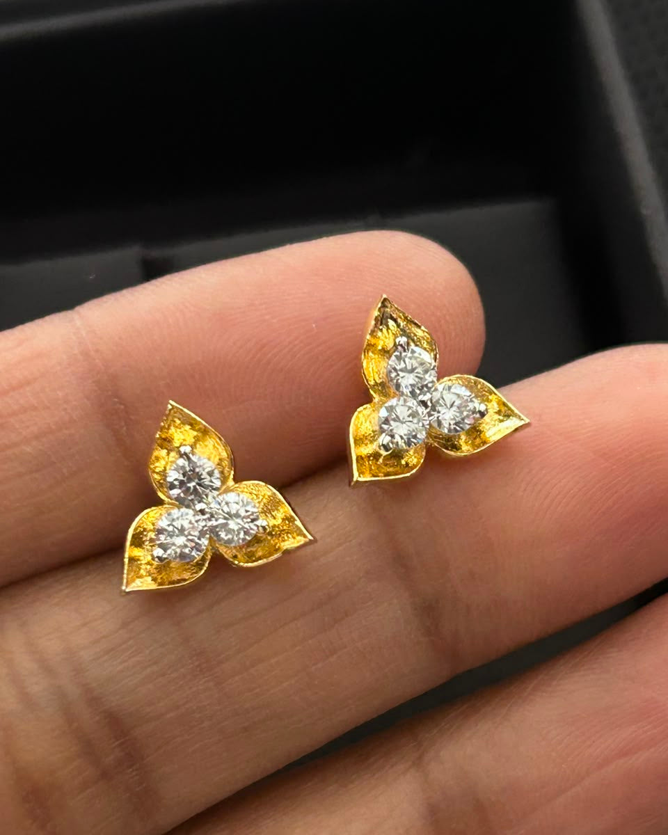 A pair of gold butterfly-shaped stud earrings with clear gemstones, held on a person's fingers against a black jewelry box background.