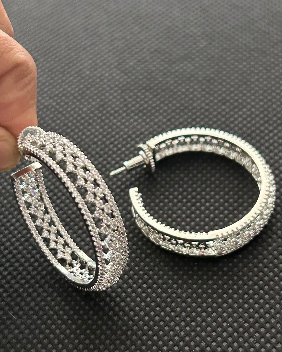 A pair of large silver-tone hoop earrings covered in clear rhinestones, with one earring held by a hand and the other resting on a dark textured surface.