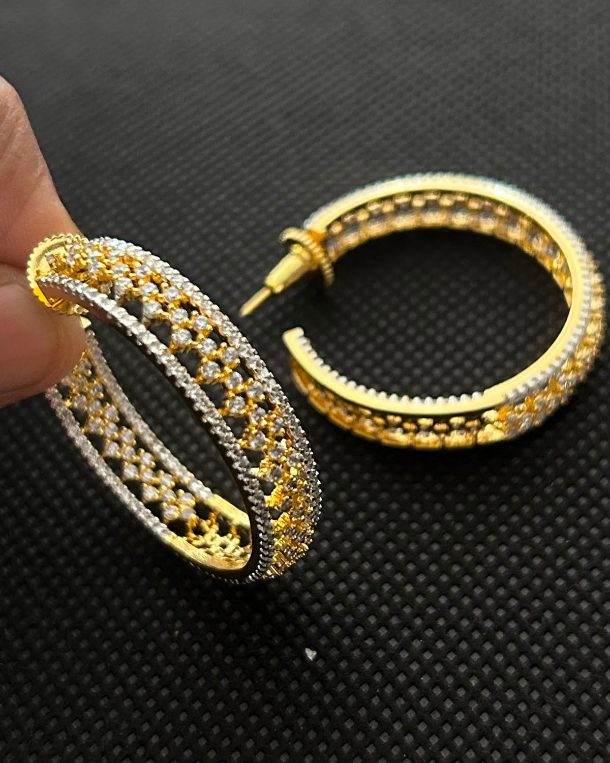 A pair of gold hoop earrings with intricate pave-set details, shown with one earring held by a hand and the other resting on a textured black background.