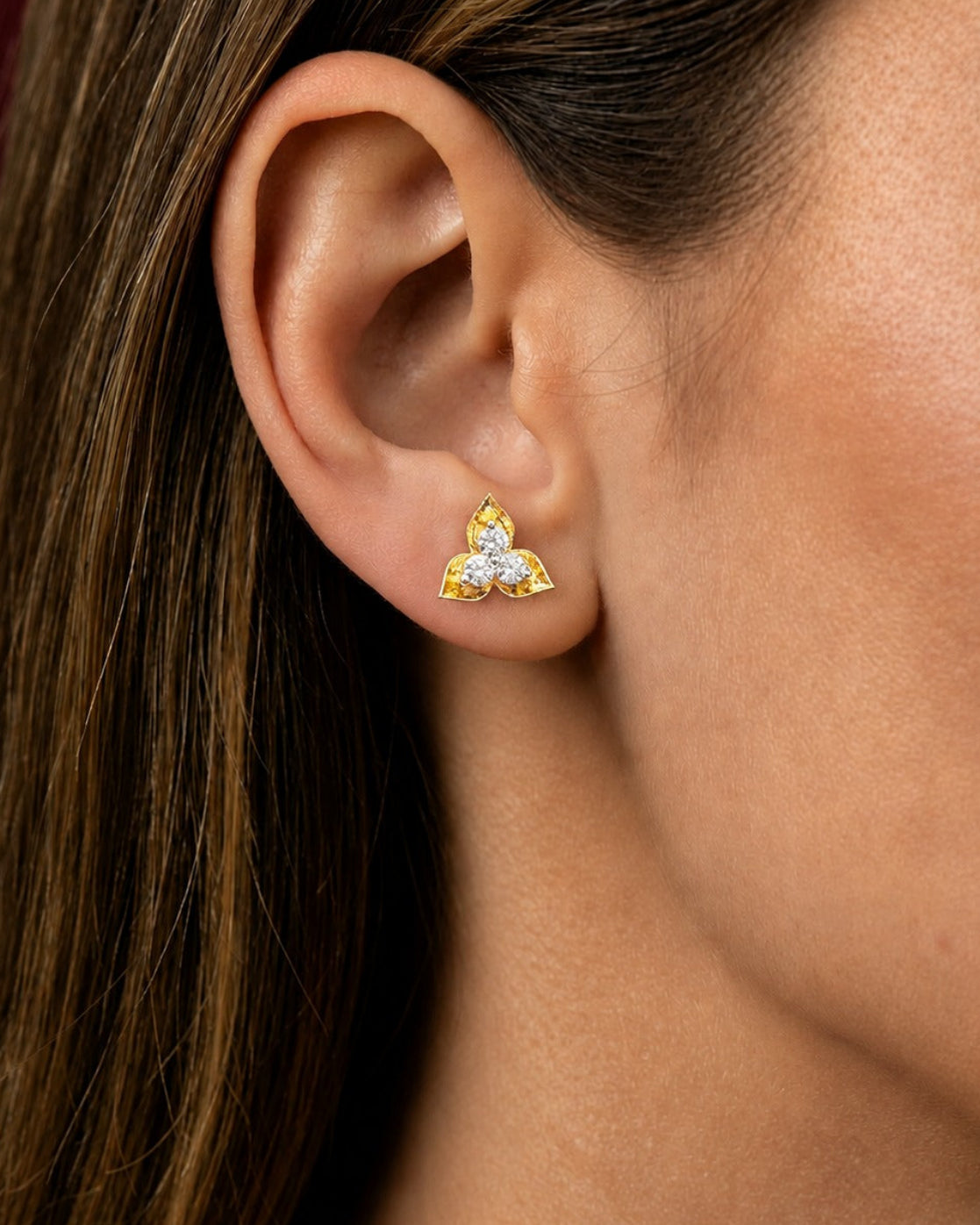 A close-up of a woman's ear wearing a small gold stud earring with three clear gemstones in a triangular arrangement.