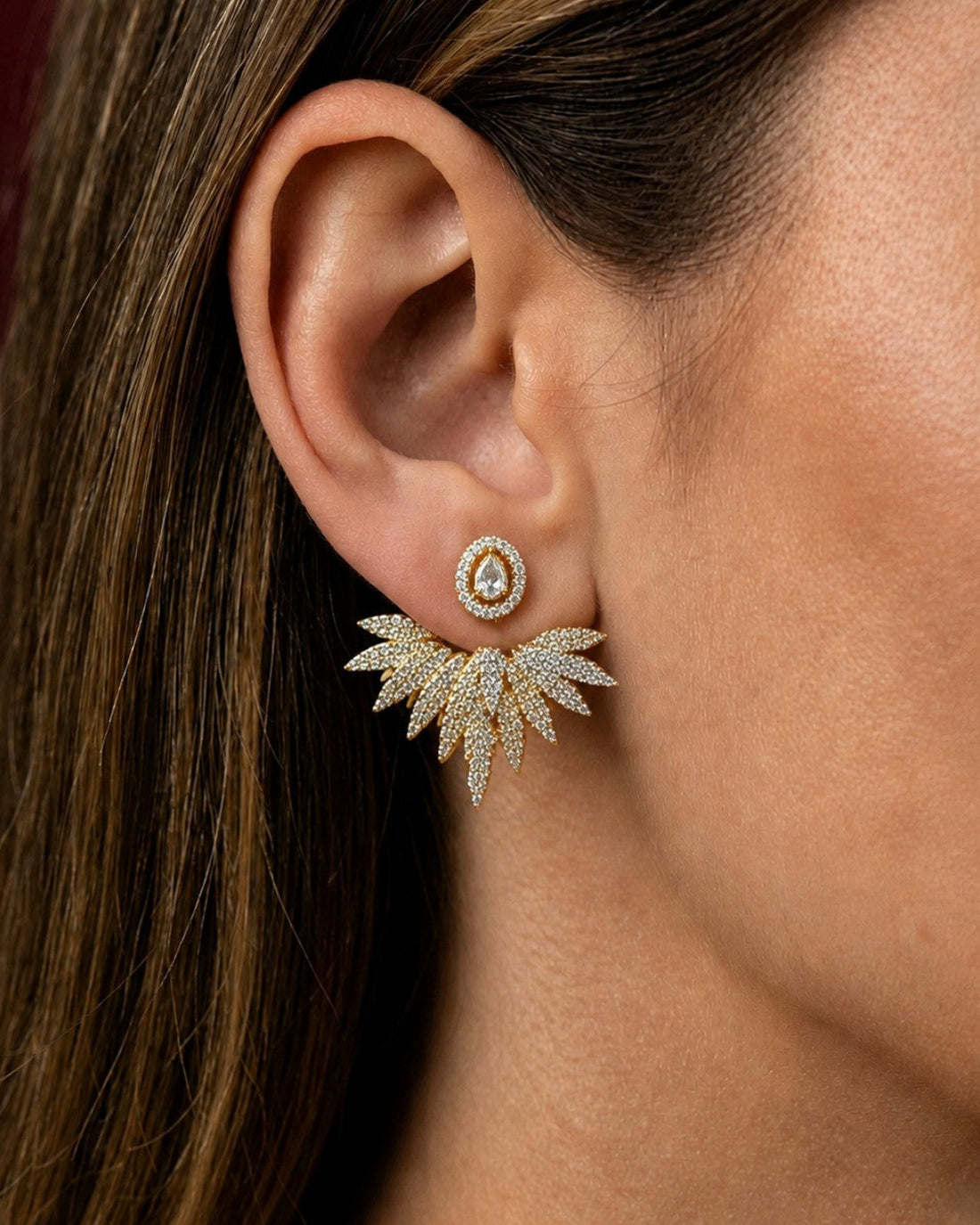 A close-up of a woman's ear wearing a gold-toned leaf-shaped stud earring with a small circular crystal cluster above it.
