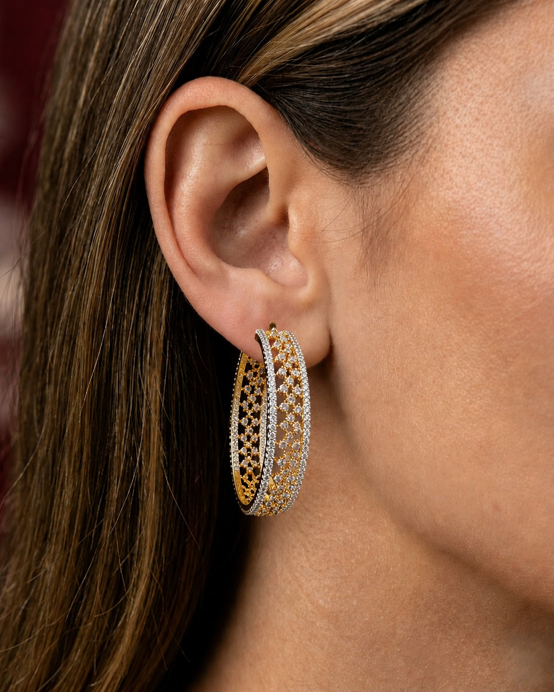 A close-up of a model's ear wearing a pair of large, intricate hoop earrings. The earrings feature a gold-tone outer hoop with an openwork filigree pattern, a silver-tone hoop paved with clear stones, and a thin gold-tone inner accent hoop.
