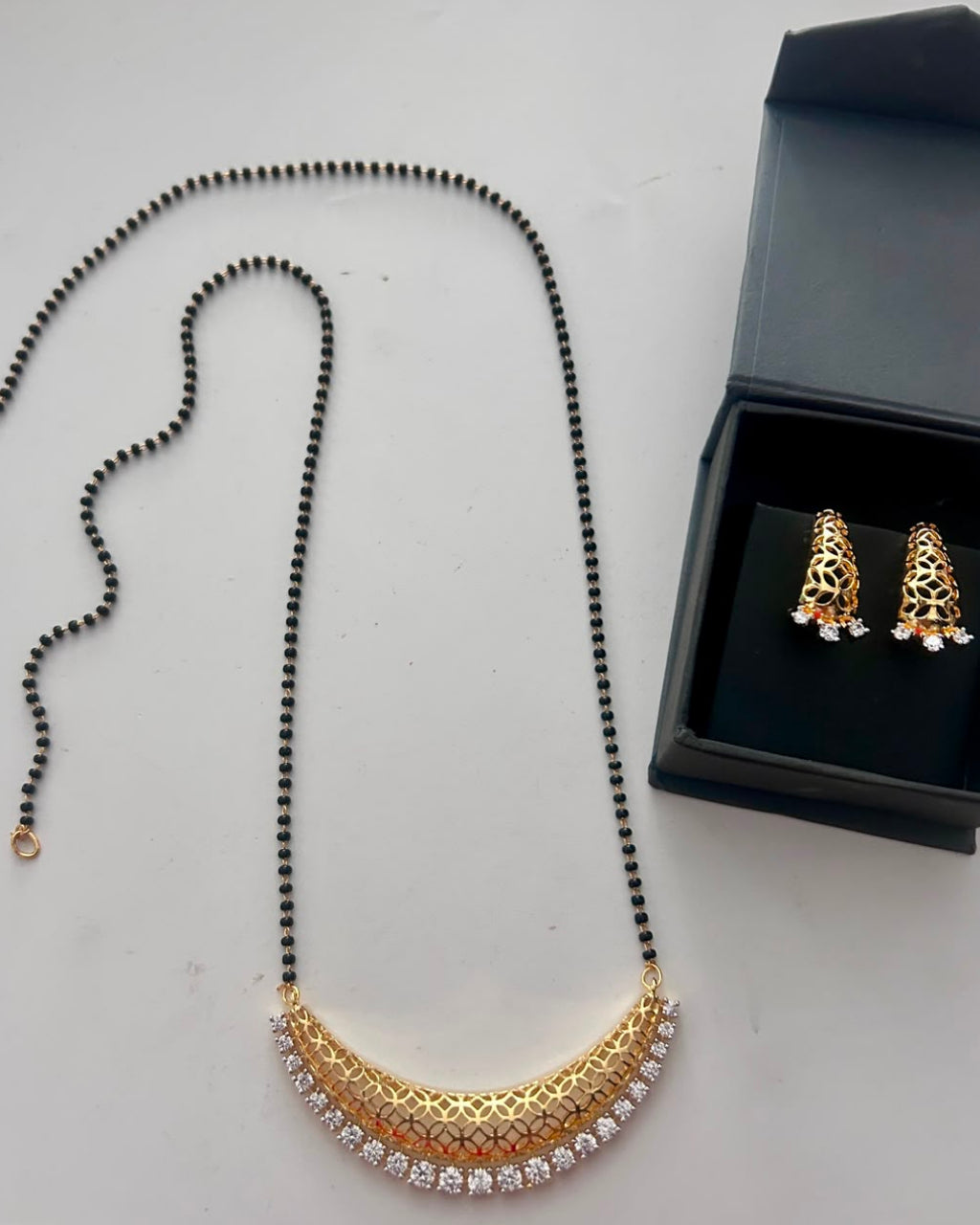 A gold crescent necklace with a black beaded upper section and clear stones along the bottom, displayed next to a black jewelry box containing matching gold earrings with clear stones.
