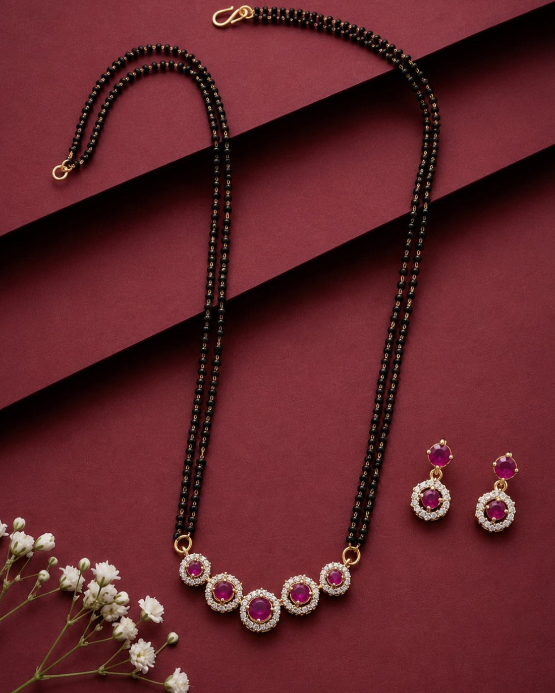 A black beaded necklace with a matching pair of drop earrings, styled on a dark red background with small white flowers. The necklace and earrings feature gold-tone settings with circular pink gemstones surrounded by clear stones.