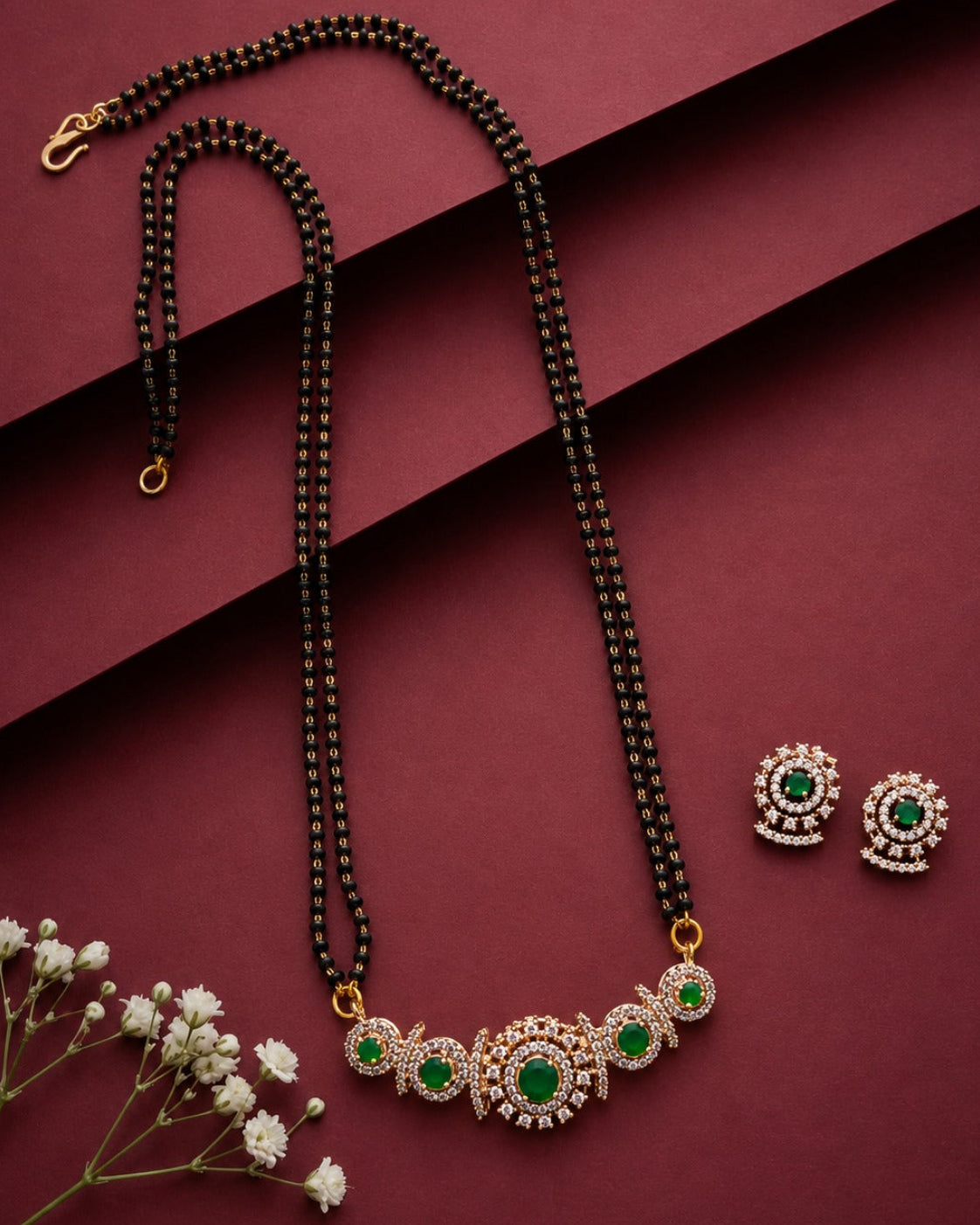 A black beaded necklace with a gold-tone and green stone pendant, displayed with matching stud earrings on a dark burgundy background with small white flowers.