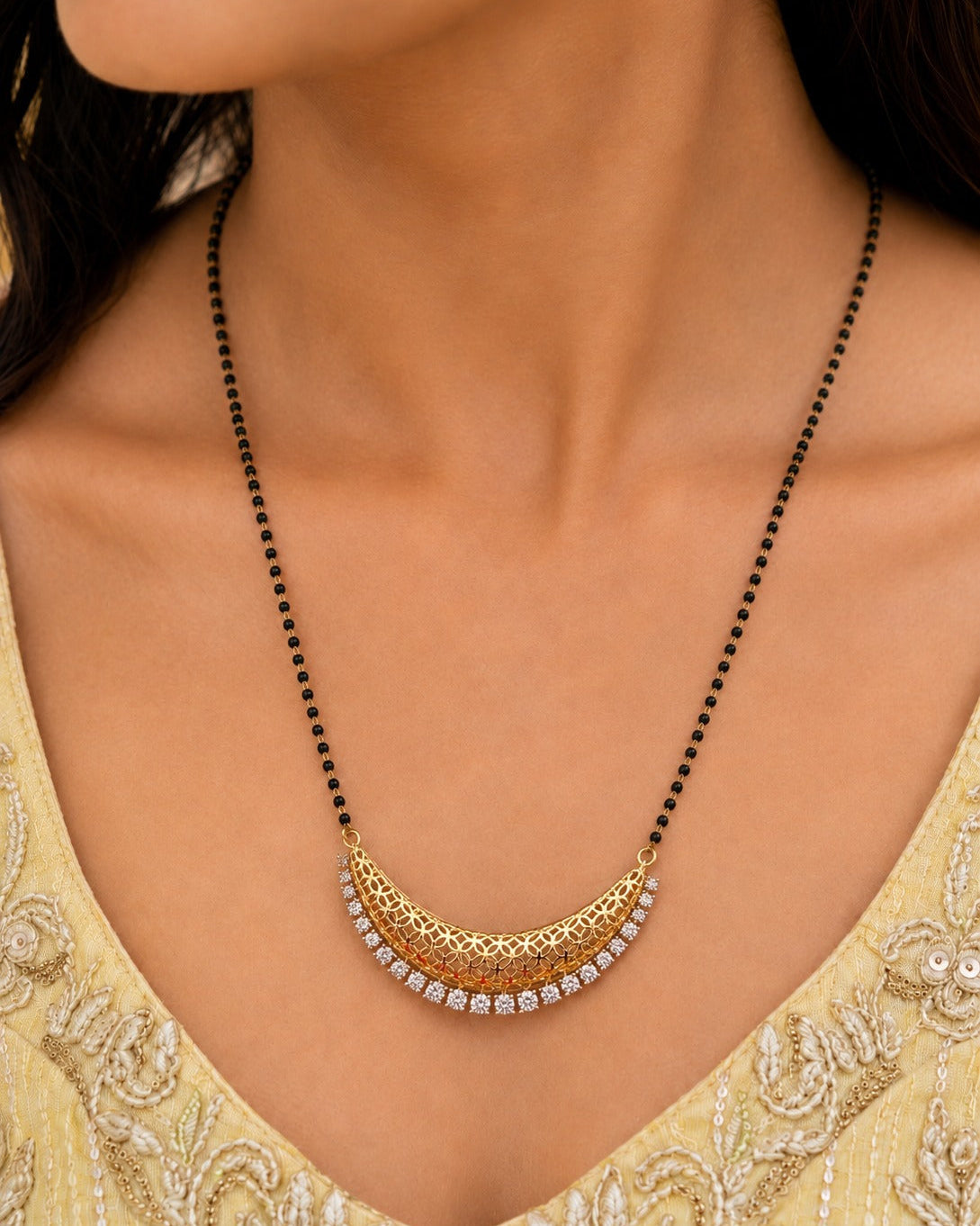 A close-up of a woman wearing a gold crescent-shaped necklace with a black beaded chain and a row of clear stones at the bottom, paired with a light yellow embroidered top.