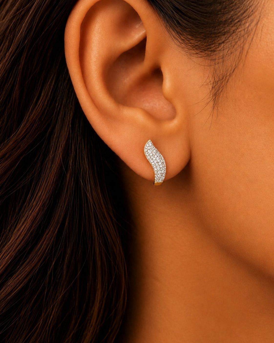 A close-up of a woman's ear wearing a small curved stud earring set with clear stones in a yellow metal setting.