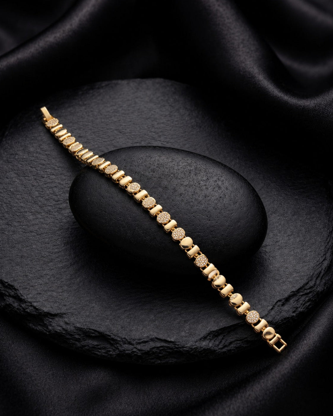 A gold tennis bracelet with small round-cut stones, displayed diagonally on a black stone slab over a draped black satin fabric background.