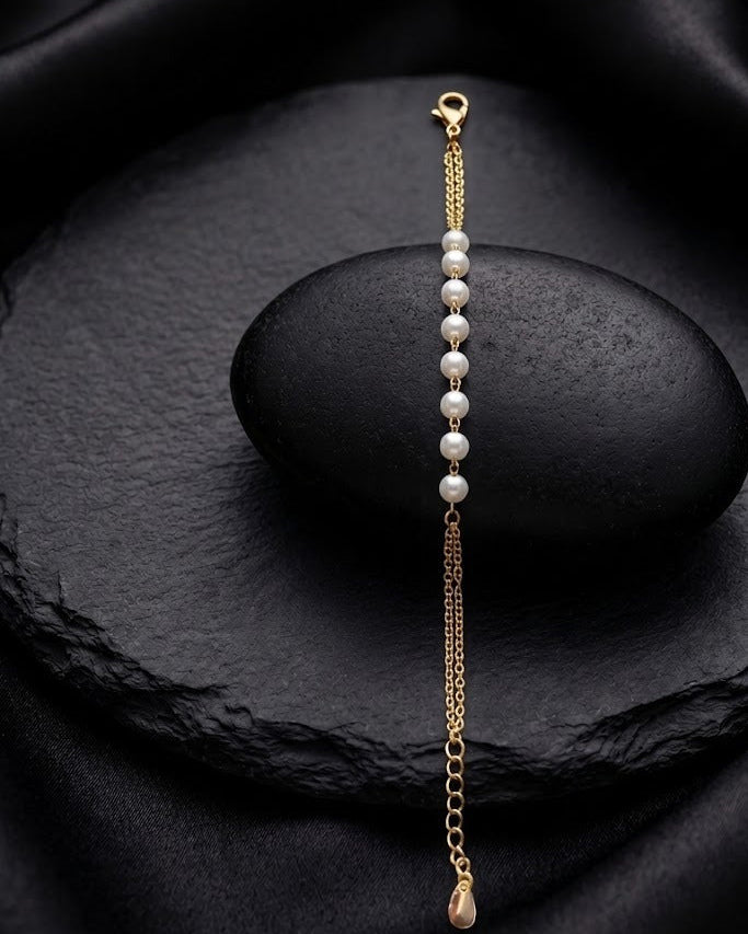 A delicate gold chain bracelet with a row of white pearls and a small gold teardrop charm, displayed on a dark stone against a black satin background.