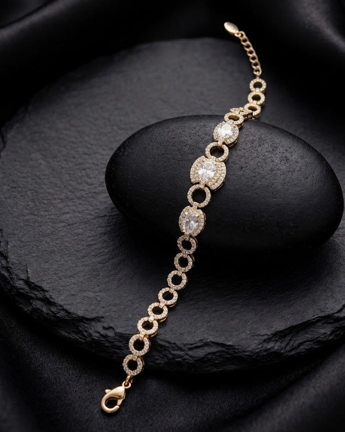 A gold chain bracelet with oval and round pavé-set clear stones, displayed on a black stone over a black satin fabric background.