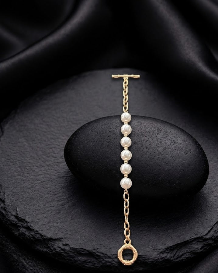A delicate gold chain bracelet with five white pearl beads and a small gold ring charm, displayed on a black stone against a black satin background.