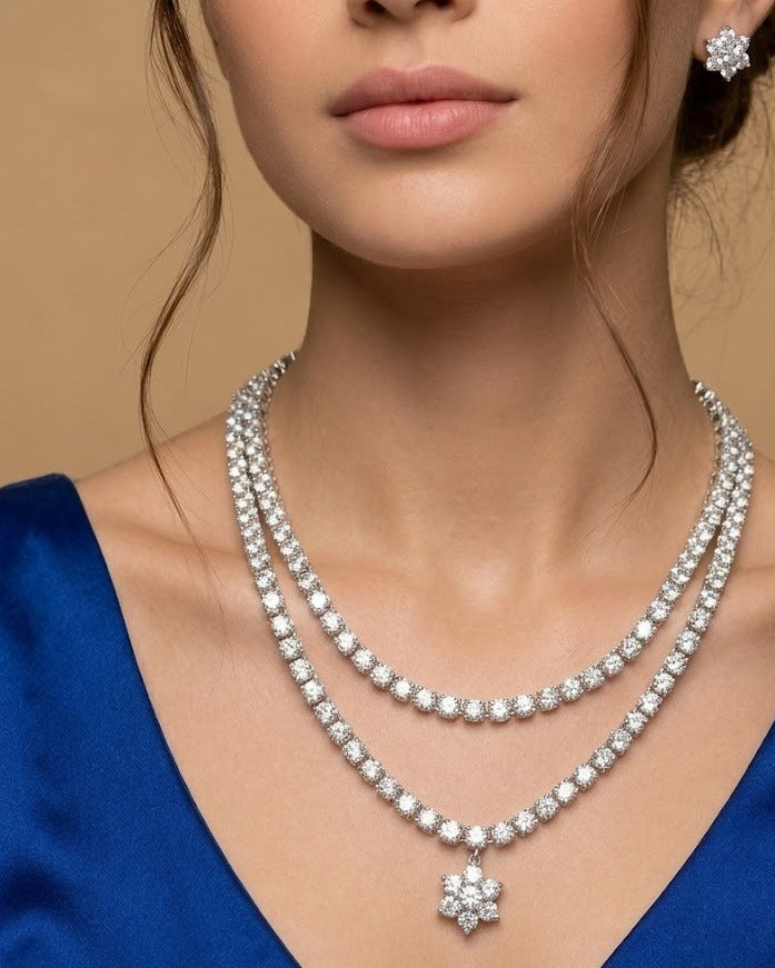 A close-up of a model wearing a double-strand diamond necklace with a flower-shaped pendant and a matching diamond stud earring, set against a warm beige background.