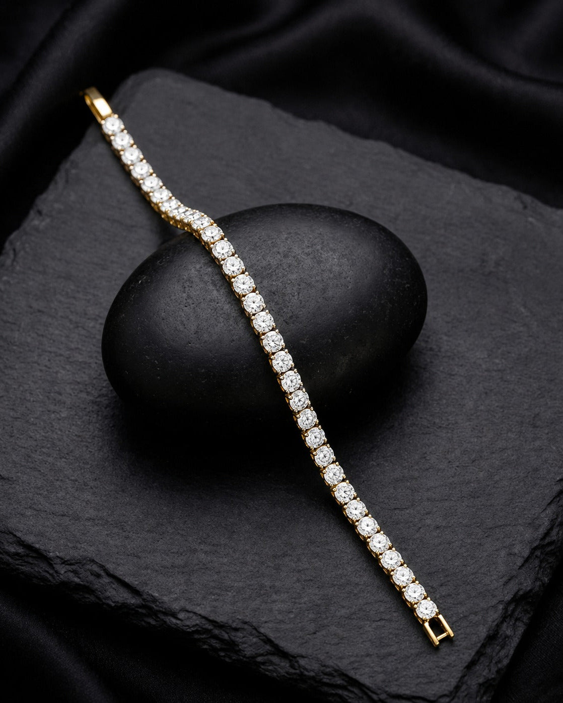 A gold tennis bracelet paved with clear round-cut gemstones, draped diagonally across a black stone on a black fabric background.