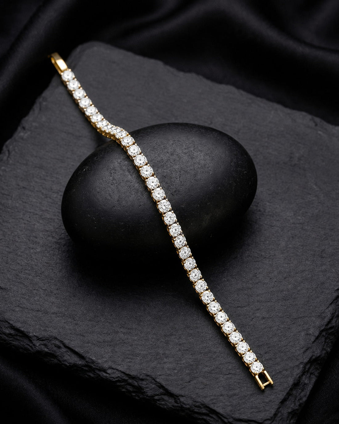 A gold tennis bracelet paved with clear round-cut gemstones, draped diagonally across a black stone on a black fabric background.