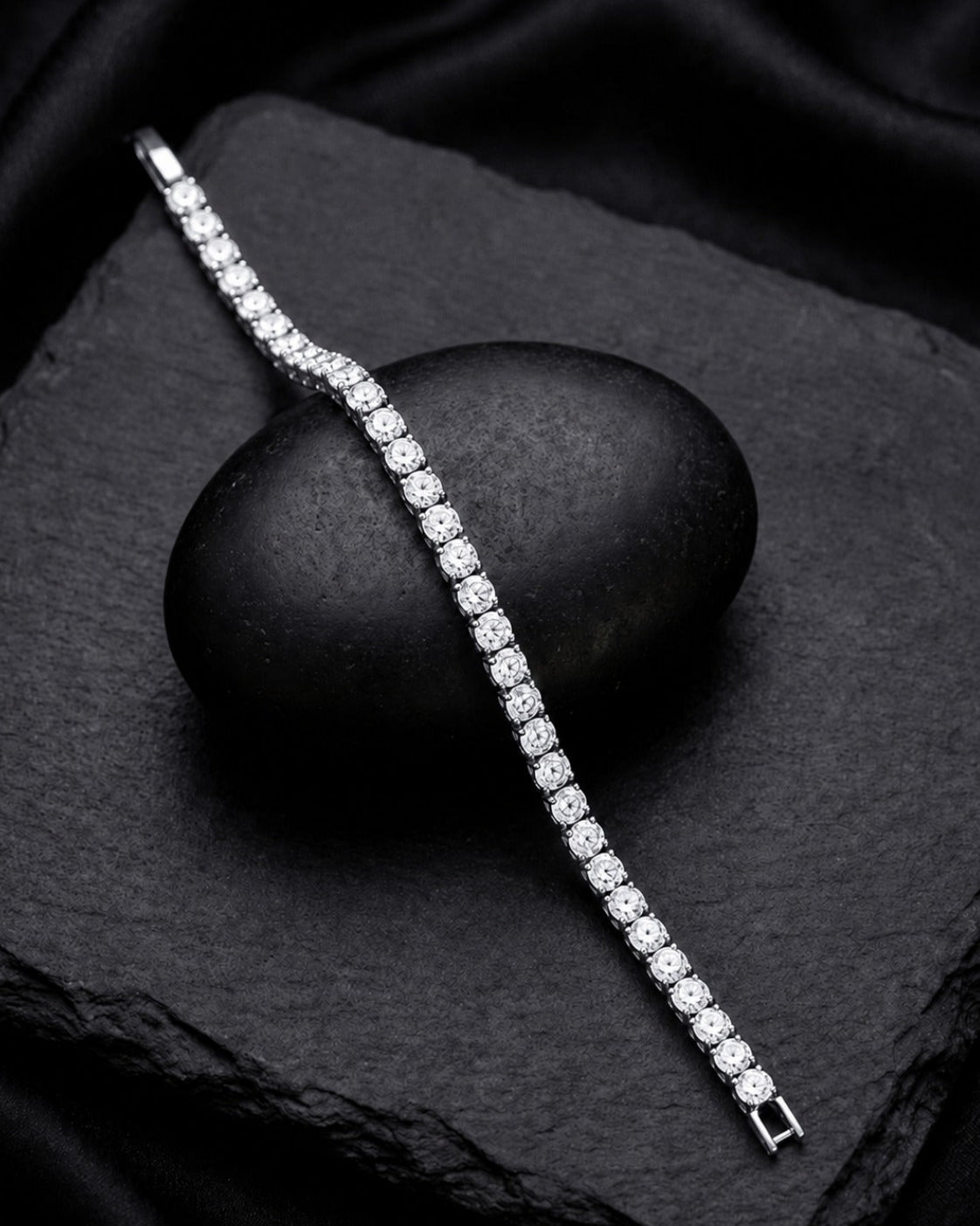 A silver tennis bracelet set with clear round-cut gemstones, elegantly draped over a smooth black stone on a textured black slate background.