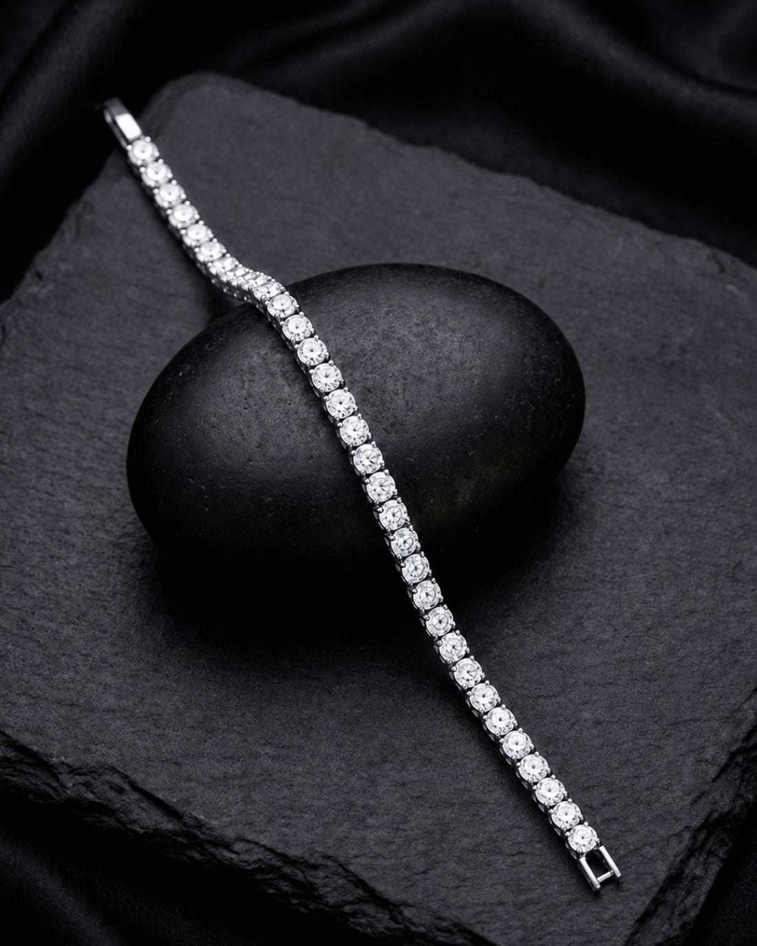 A silver tennis bracelet set with clear round-cut gemstones, elegantly draped over a smooth black stone on a textured black slate background.